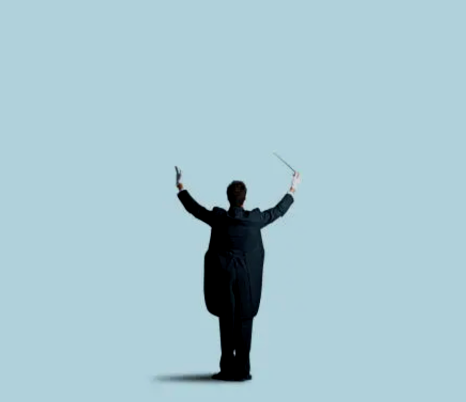 A conductor holding a baton in front of sheet music on a stand, with a wooden surface and blue background.