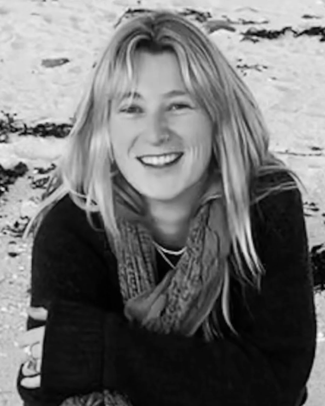 A smiling woman with long hair sitting on a beach with sand and rocks in the background.