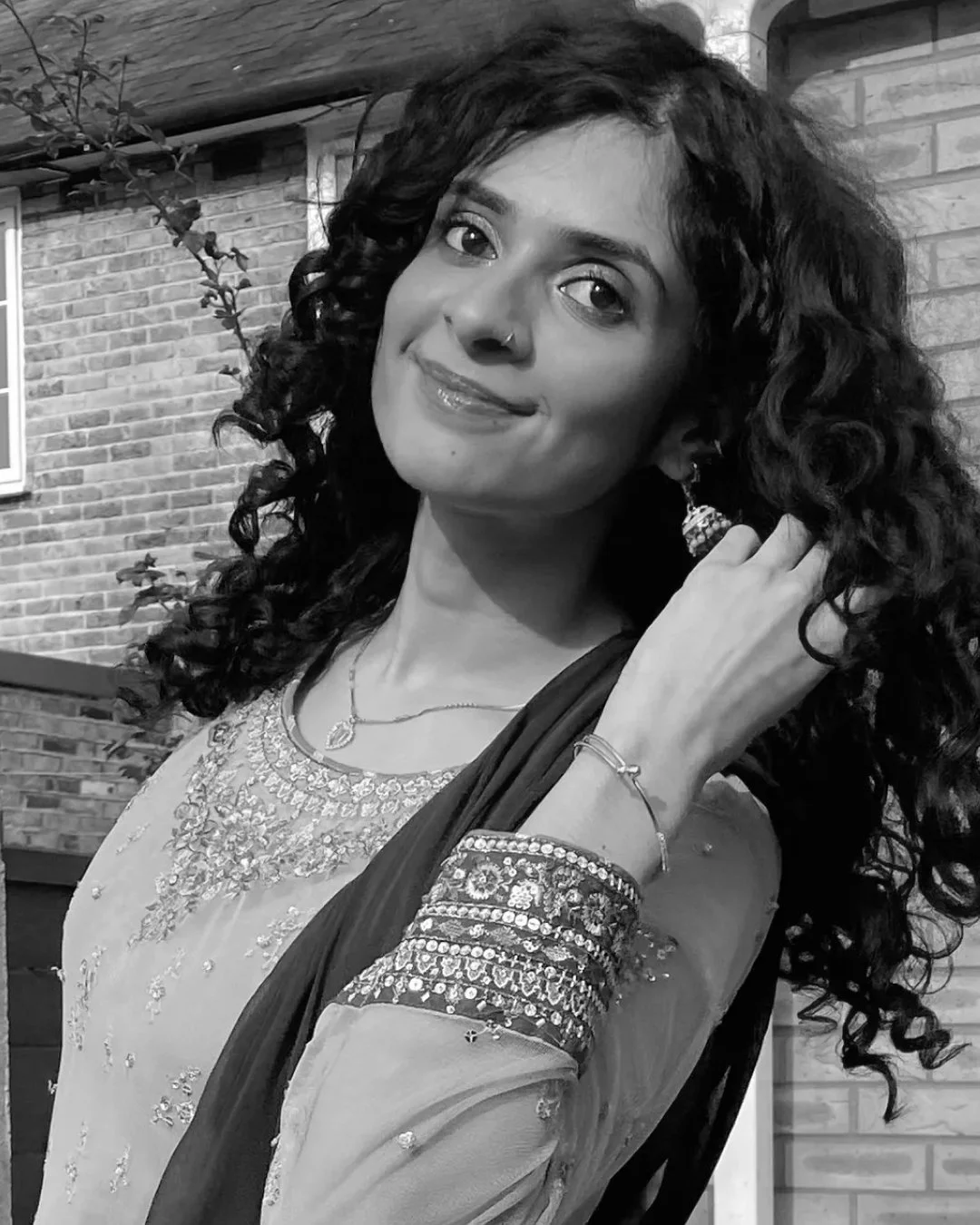 A woman with curly hair, wearing traditional attire with jewelry, standing in front of a brick wall, smiling and adjusting her earring.