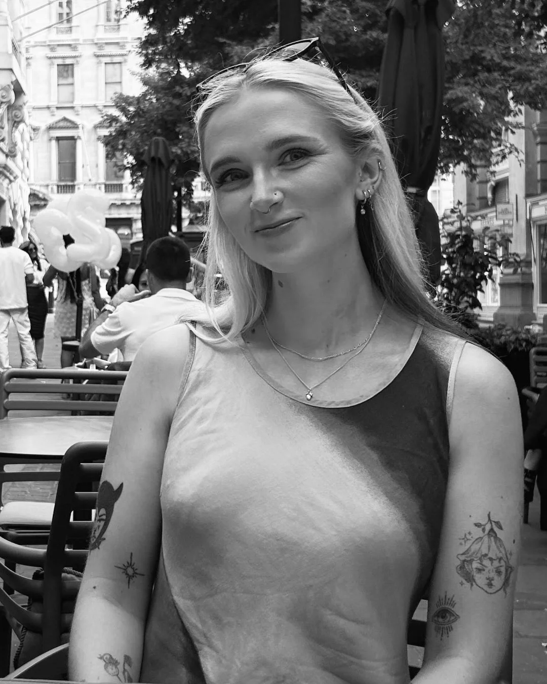 A young woman with tattoos on her arms, wearing a sleeveless top, sitting outdoors at a cafe. She has long blonde hair, earrings, and layered necklaces. In the background, there are other people, trees, and buildings, in an urban setting.