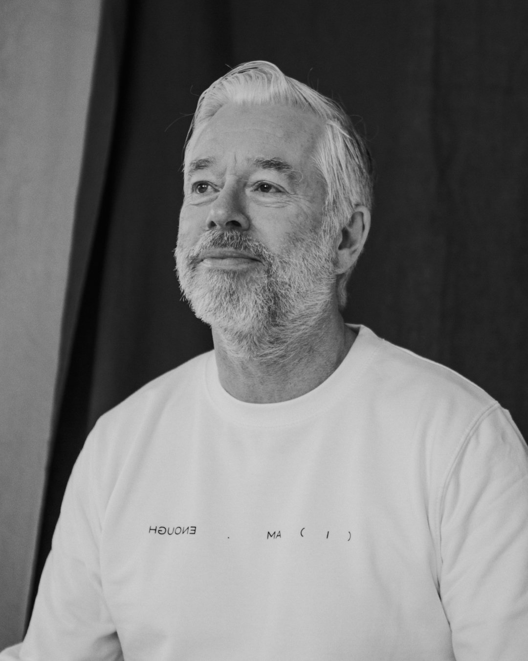 Black and white photo of a middle-aged man with a beard and mustache, looking slightly upward, wearing a white T-shirt, with a background of a dark wall and doorway.