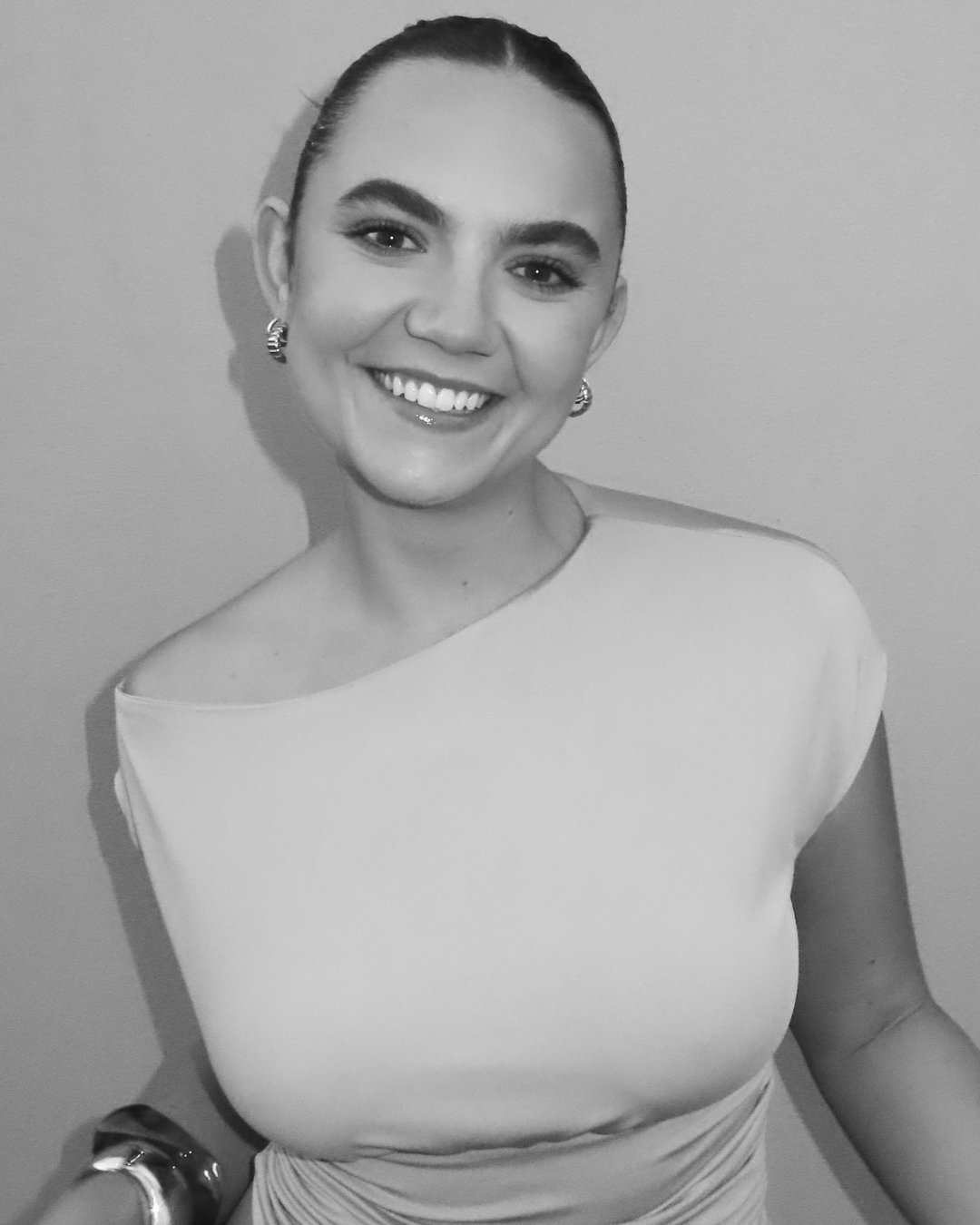 Black-and-white portrait of a smiling woman with short hair, wearing earrings, a one-shoulder top, and a wristwatch, standing against a plain background.