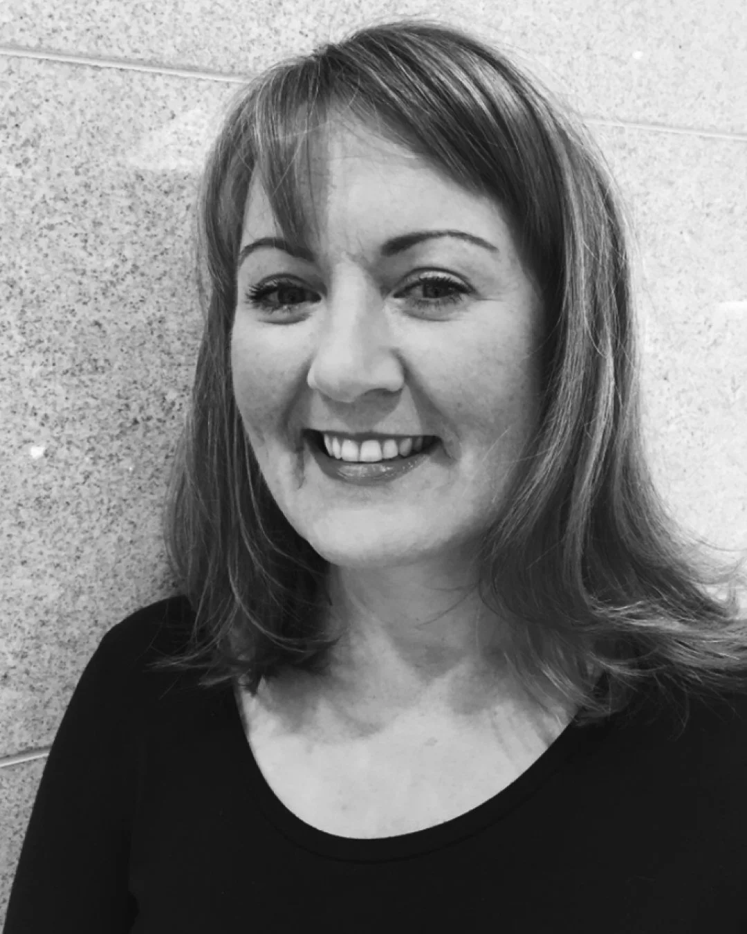 Black and white portrait of a smiling woman with shoulder-length hair, wearing a black top, standing against a textured wall.