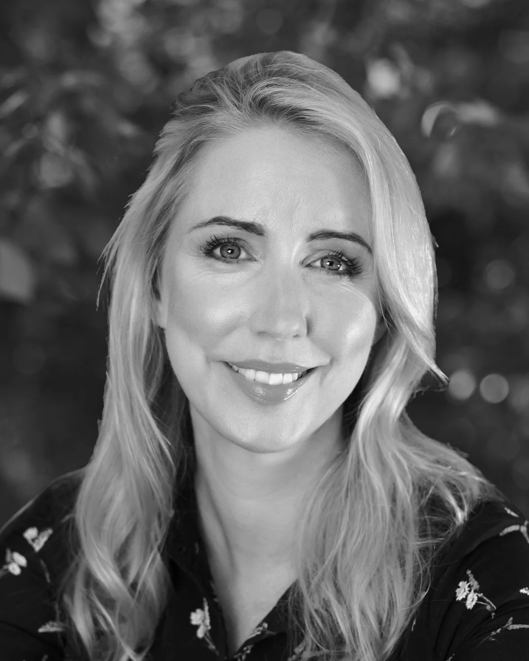 Portrait of a smiling woman with light-colored hair and eyes, wearing a dark top with a floral pattern, outdoors with a blurred natural background.