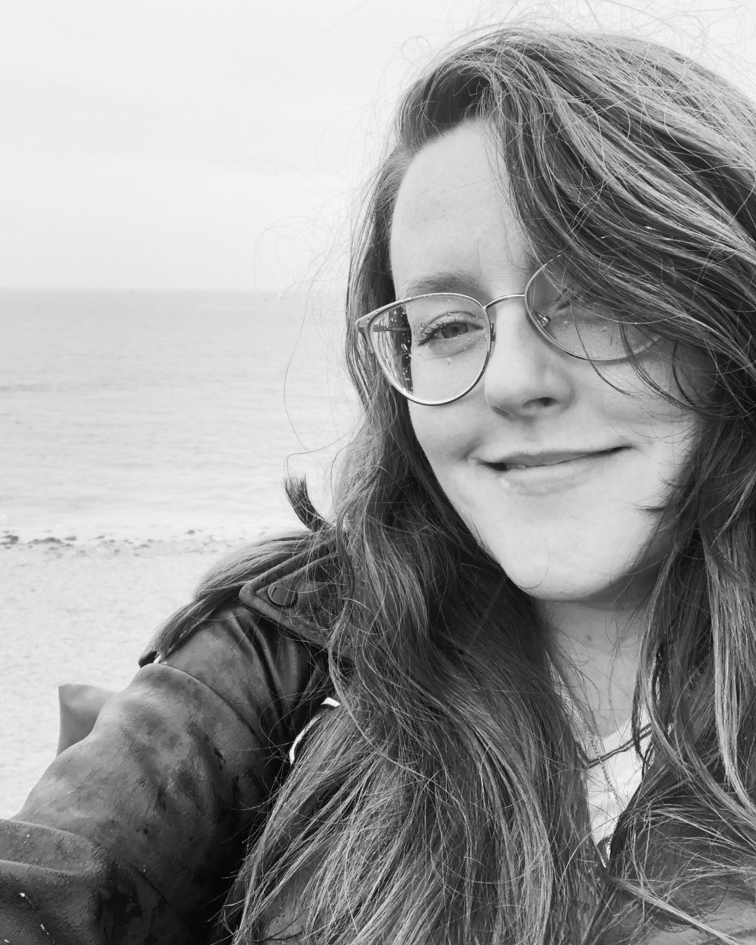A woman with long wavy hair and glasses smiling at the camera at the beach.
