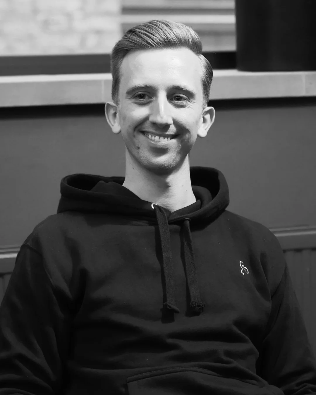 A man smiling, wearing a black hoodie, sitting indoors, black and white photo.