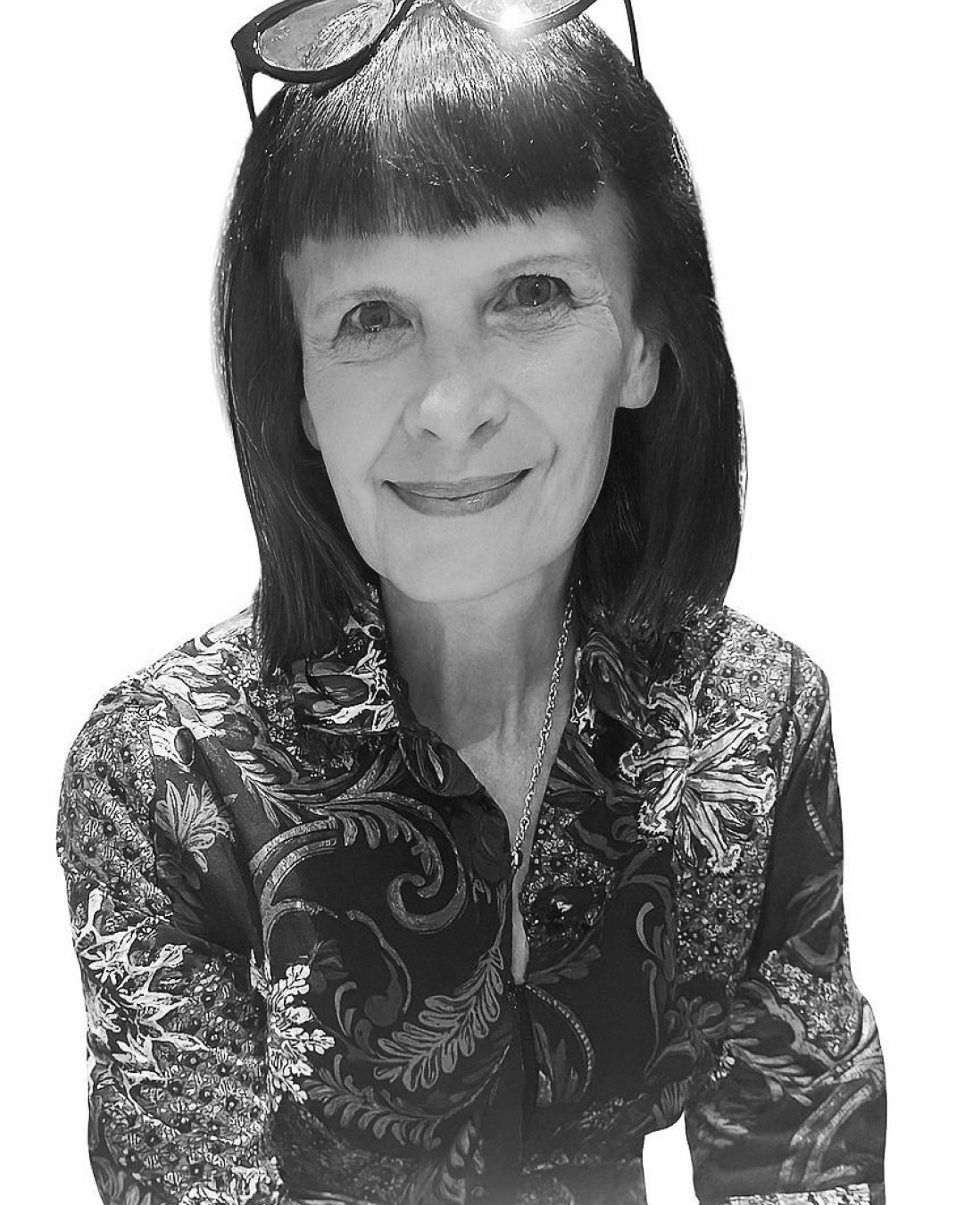 Black-and-white portrait of a middle-aged woman with dark hair and bangs, wearing glasses on her head and a patterned blouse with floral and swirling designs.