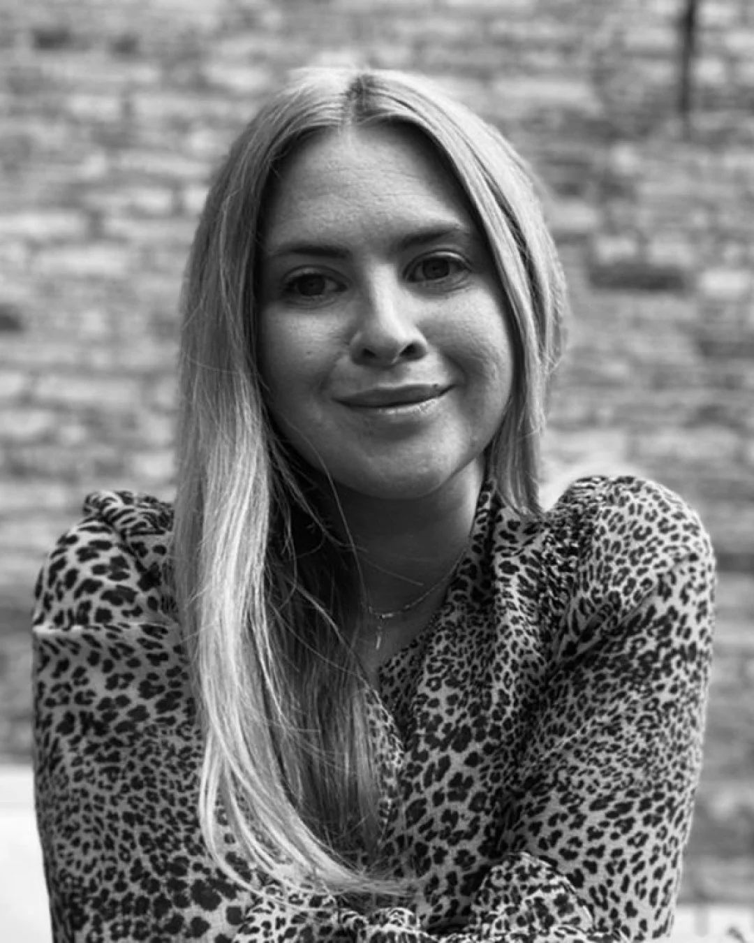 Black and white photo of a woman with long hair, smiling, wearing a leopard print top, in front of a brick wall.