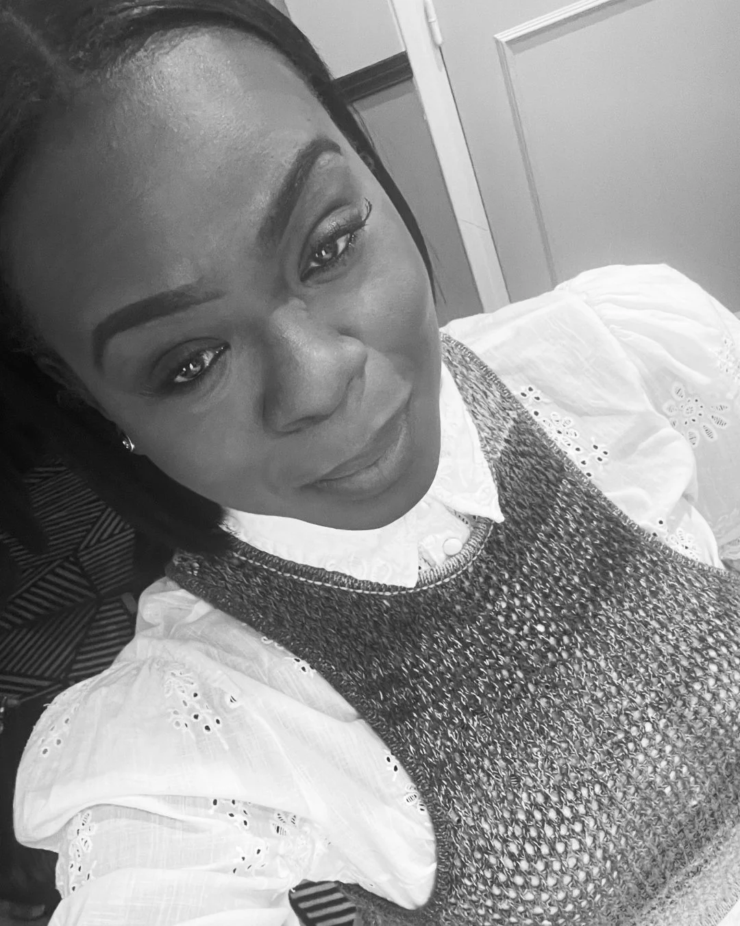 Black and white photo of a woman smiling, wearing a textured vest over a blouse with eyelet embroidery and puffed sleeves.