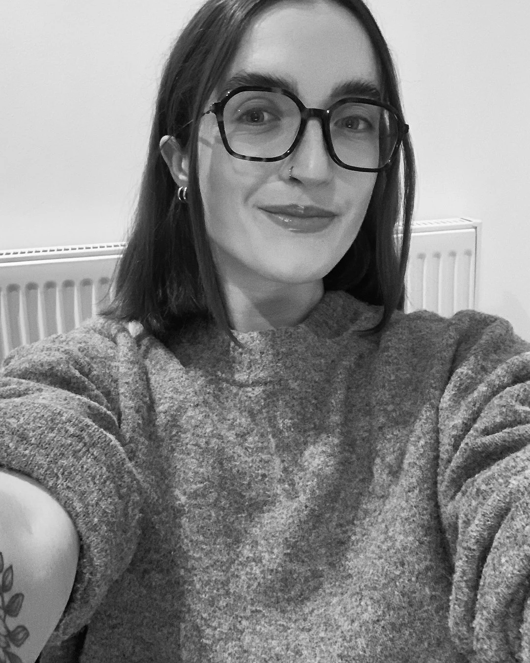 A woman taking a selfie, wearing glasses and a sweater, with a radiator in the background.