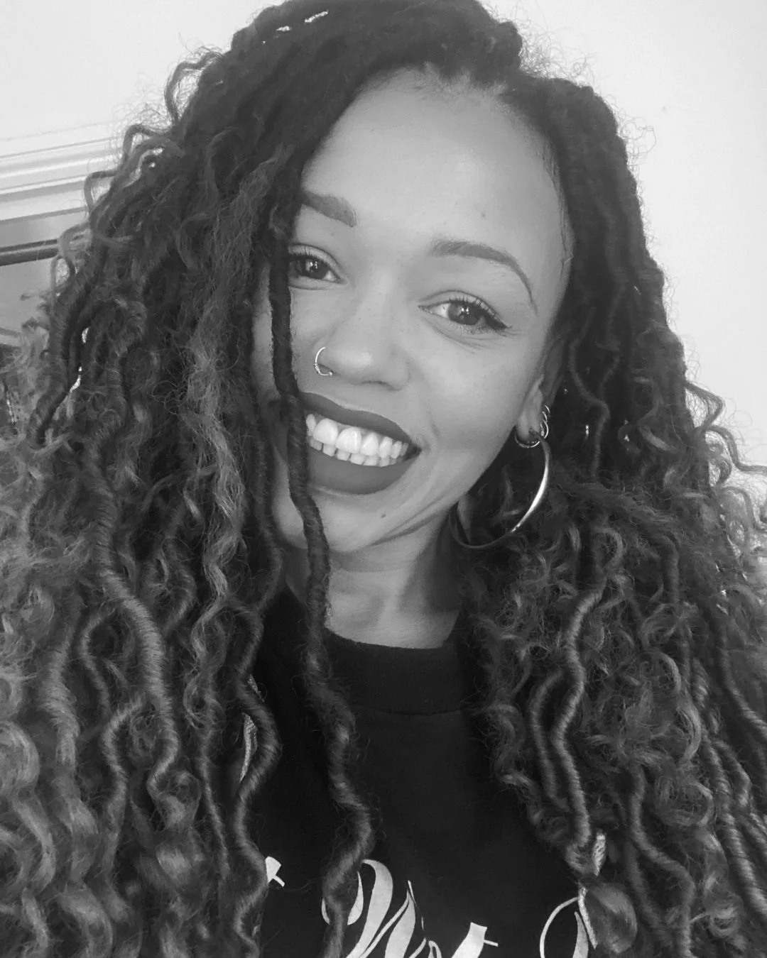 A woman with long, curly hair, wearing hoop earrings and a nose ring, smiling at the camera in black and white.
