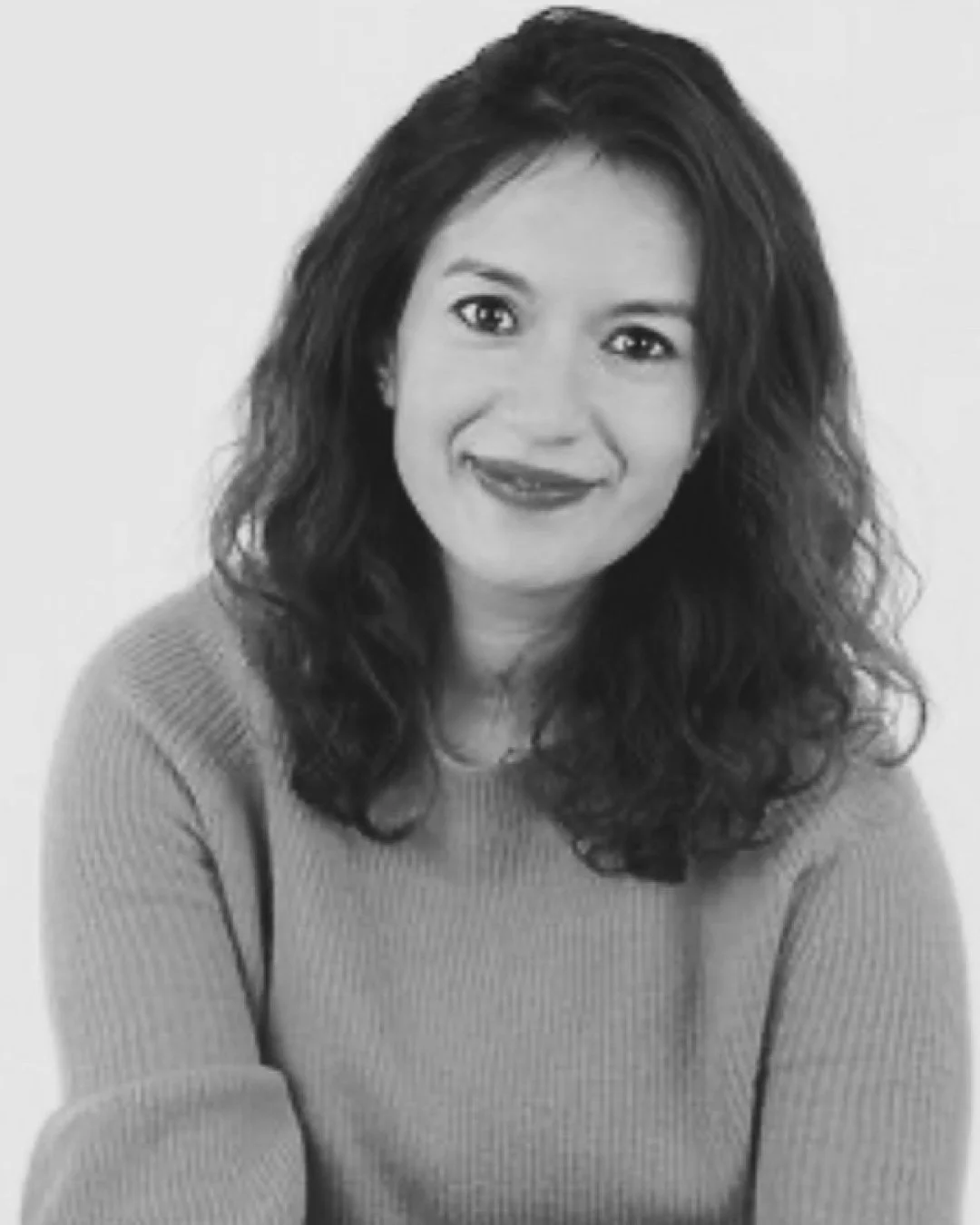 Black and white portrait of a woman with shoulder-length curly hair, wearing a long-sleeved top, smiling at the camera.