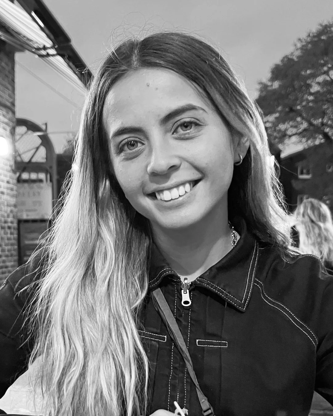 Black and white photo of a young woman with long hair, smiling, outdoors with trees and a house in the background.