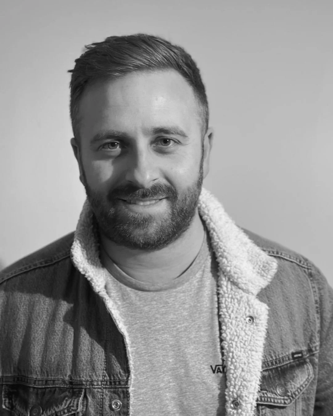 Black and white portrait of a smiling man with short hair, beard, and mustache, wearing a denim jacket with a fleece collar and a light-colored t-shirt underneath.