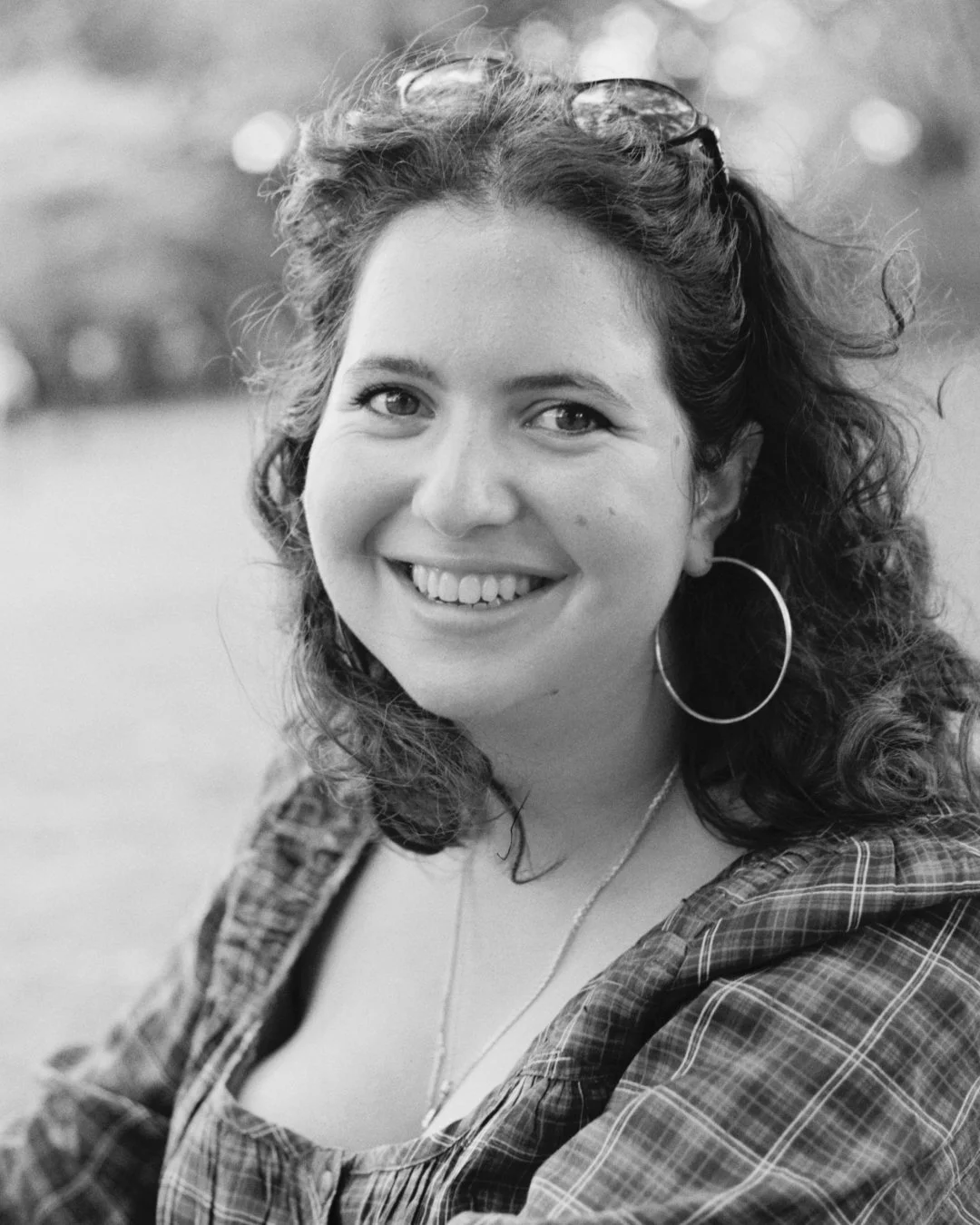 A young woman with curly hair, smiling, wearing hoop earrings, a necklace, sunglasses on her head, and a plaid shirt in an outdoor setting, black and white photograph.