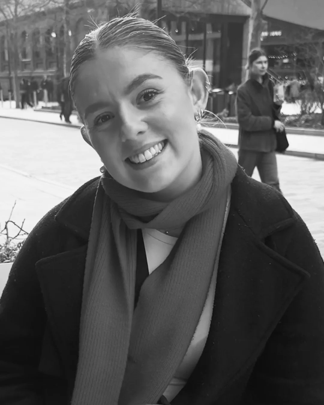 A young woman smiling at the camera outdoors with other people and buildings in the background