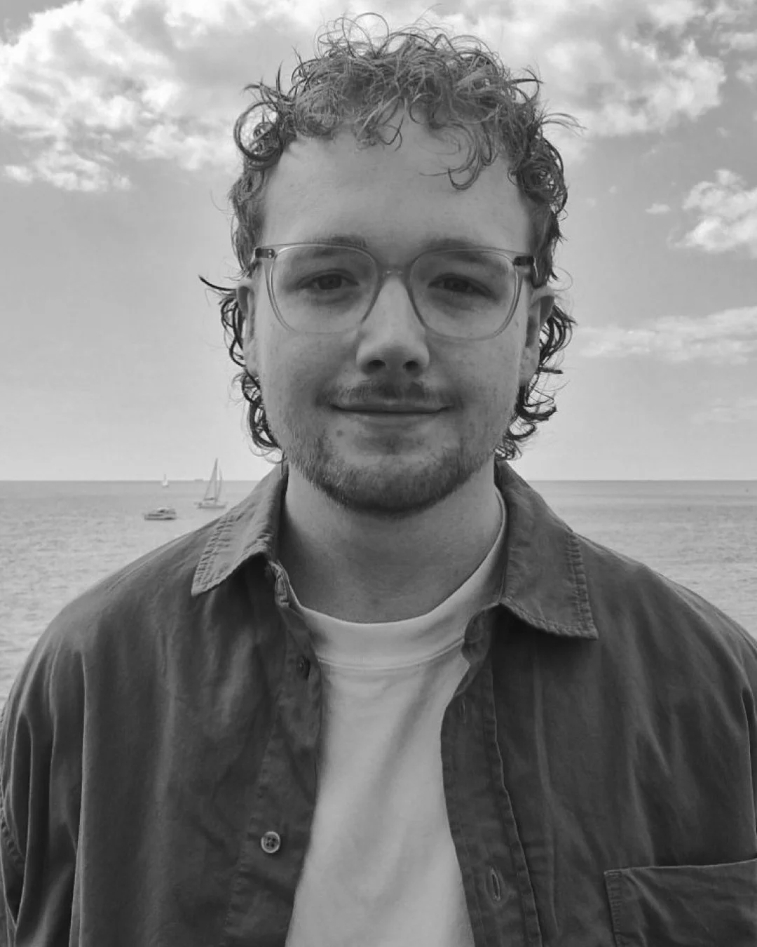 A man with curly hair and glasses smiling outdoors at the beach with sailboats and clouds in the sky.
