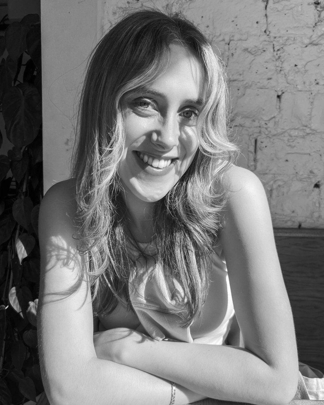 A young woman smiling with hair falling over her shoulders, seated against a brick wall, black and white photograph.