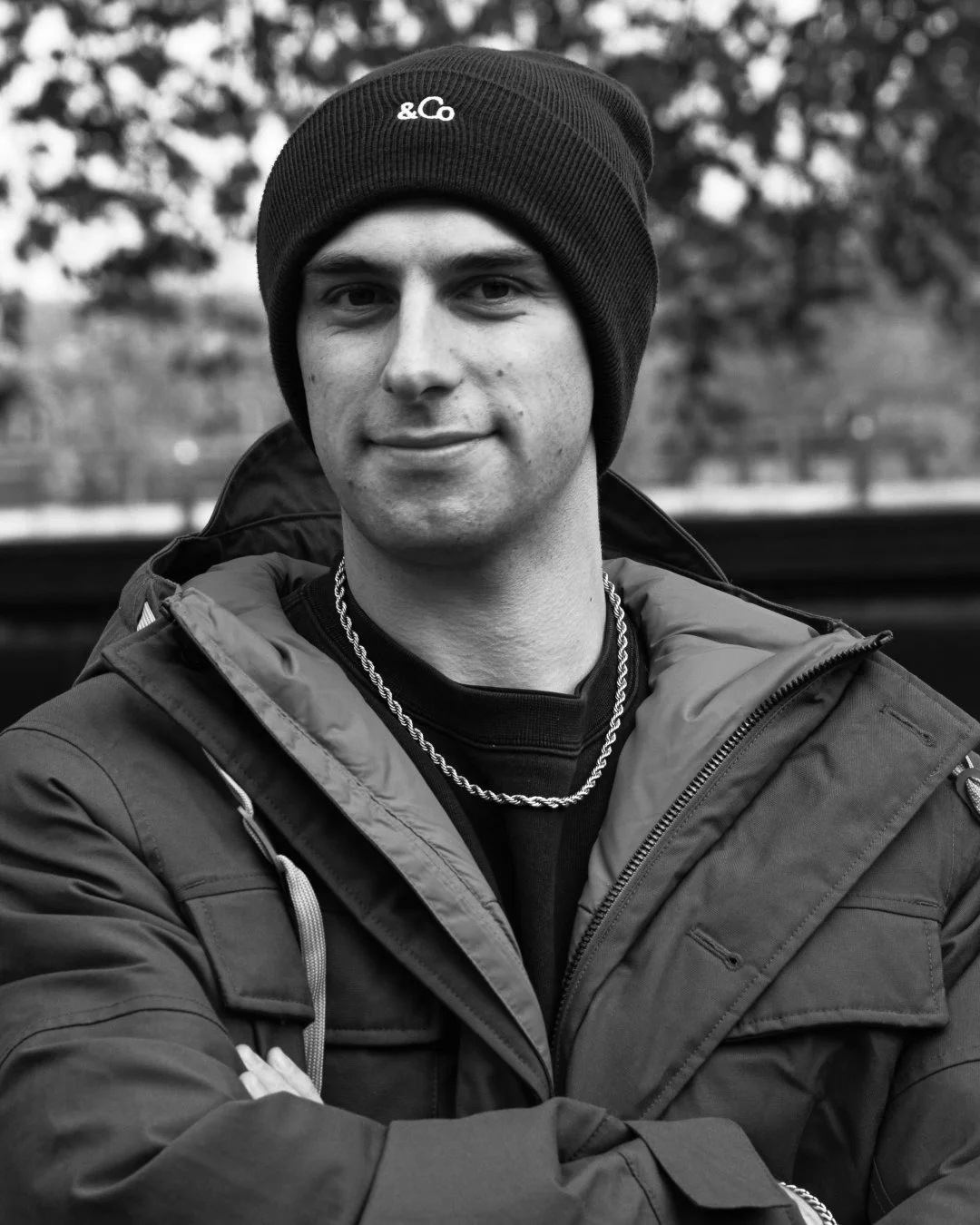 Black and white photo of a young man wearing a beanie hat, a chain necklace, and a winter jacket, standing outdoors with blurred trees in the background.