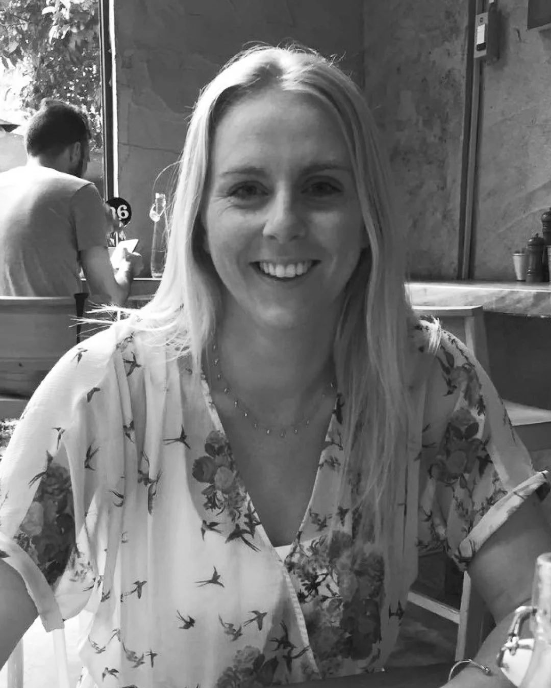 A woman with long blonde hair smiling at the camera, wearing a floral blouse and a necklace, in a restaurant setting with other patrons in the background.