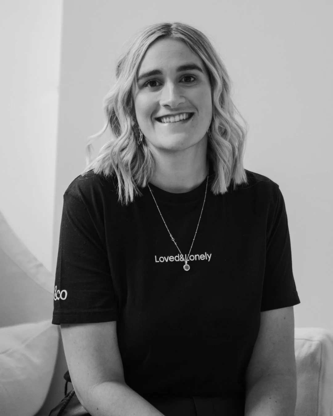 A young woman with shoulder-length wavy hair, smiling, wearing a black T-shirt that says 'Loved&lonely' and earrings.