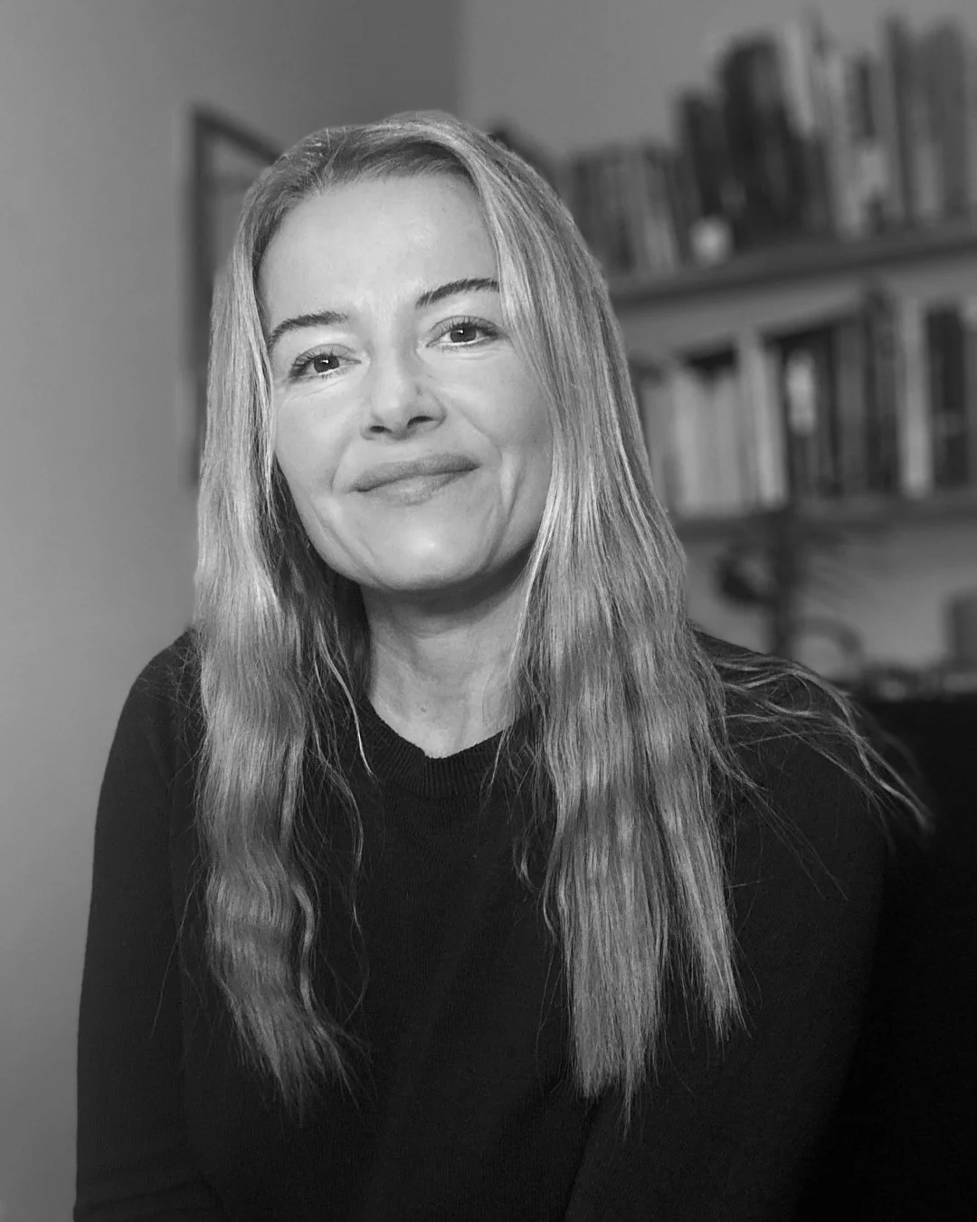 Black and white portrait of a woman with long, wavy hair, smiling gently, sitting in front of a blurred background with bookshelves.