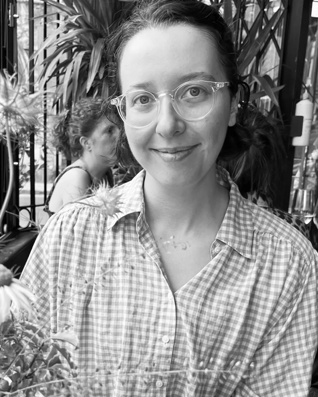 A woman with glasses smiling at camera, wearing a checkered shirt, with another woman in the background near plants.