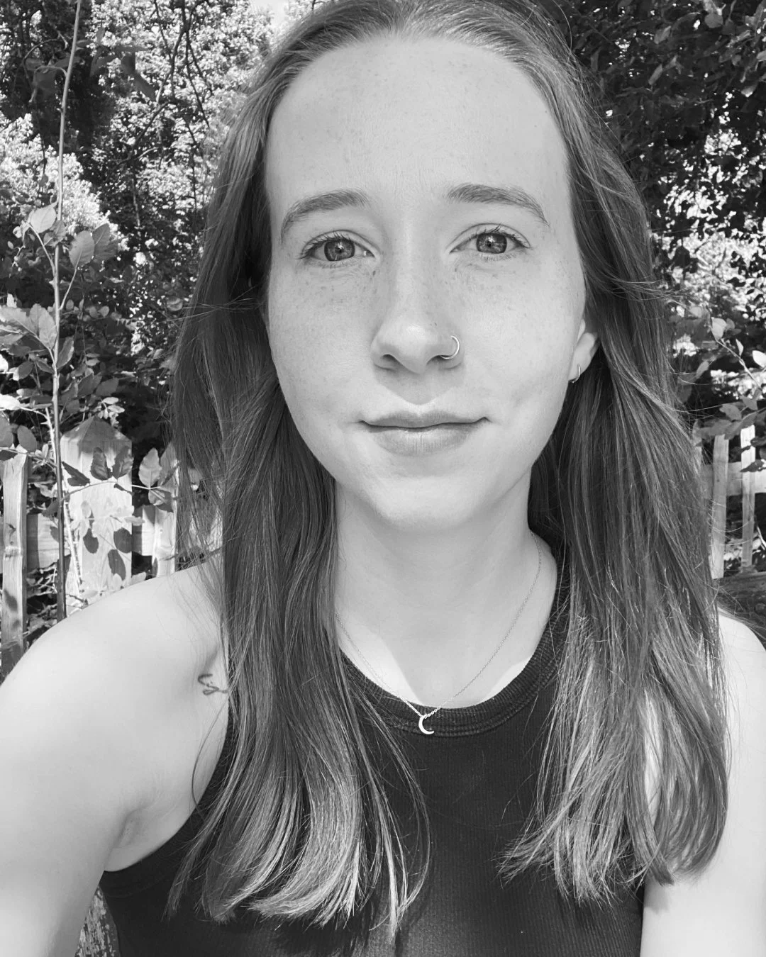 Black and white photo of a young woman with long hair, smiling slightly, wearing a sleeveless top, a small hoop nose ring, a necklace, and earrings, outdoors with trees and a fence in the background.