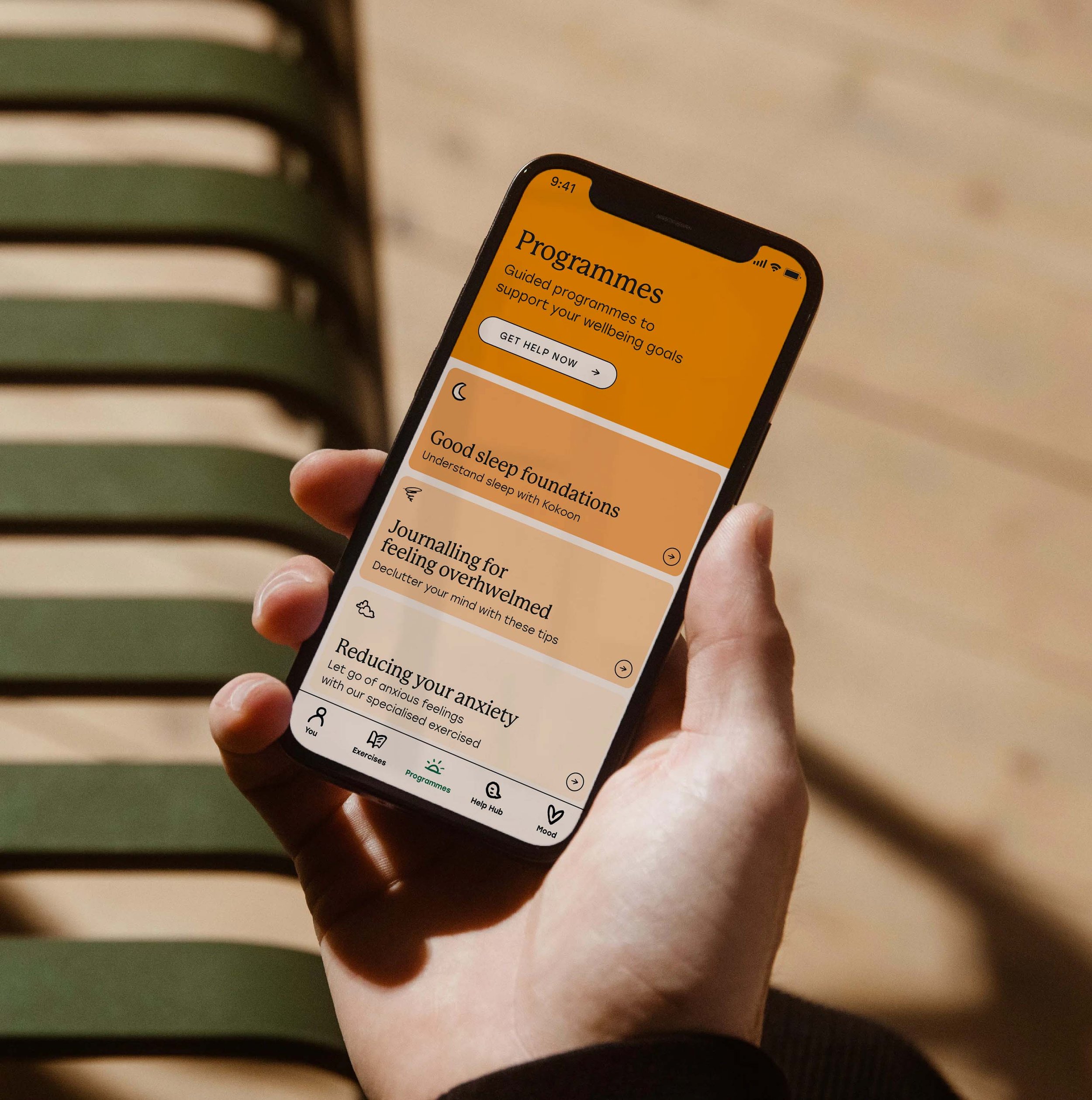 A hand holding a smartphone displaying a wellness app with sections on programmes, sleep, anxiety, mood, exercises, help hub, and more.