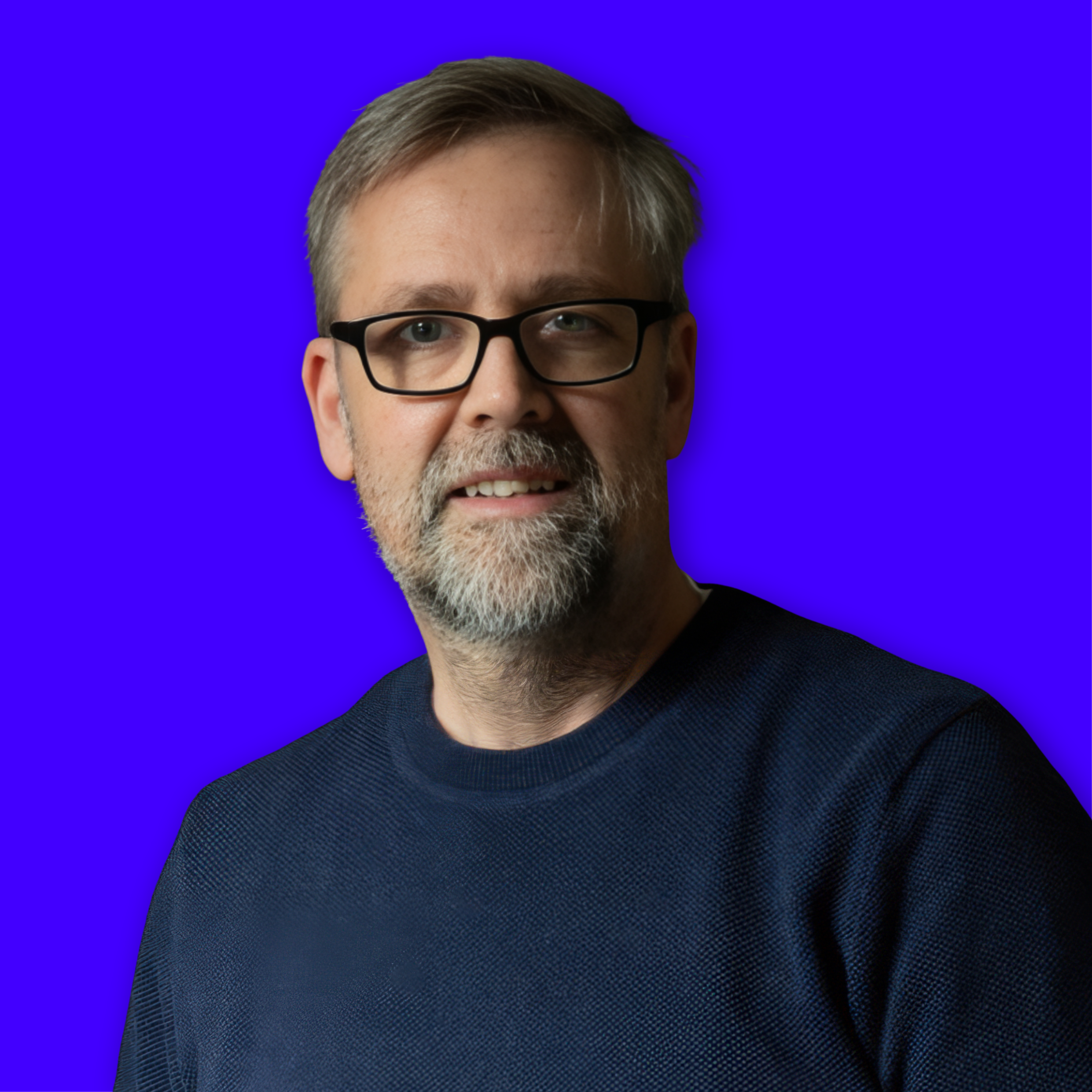 A middle-aged man with gray hair and beard, wearing glasses and a dark sweater, smiling against a solid blue background.