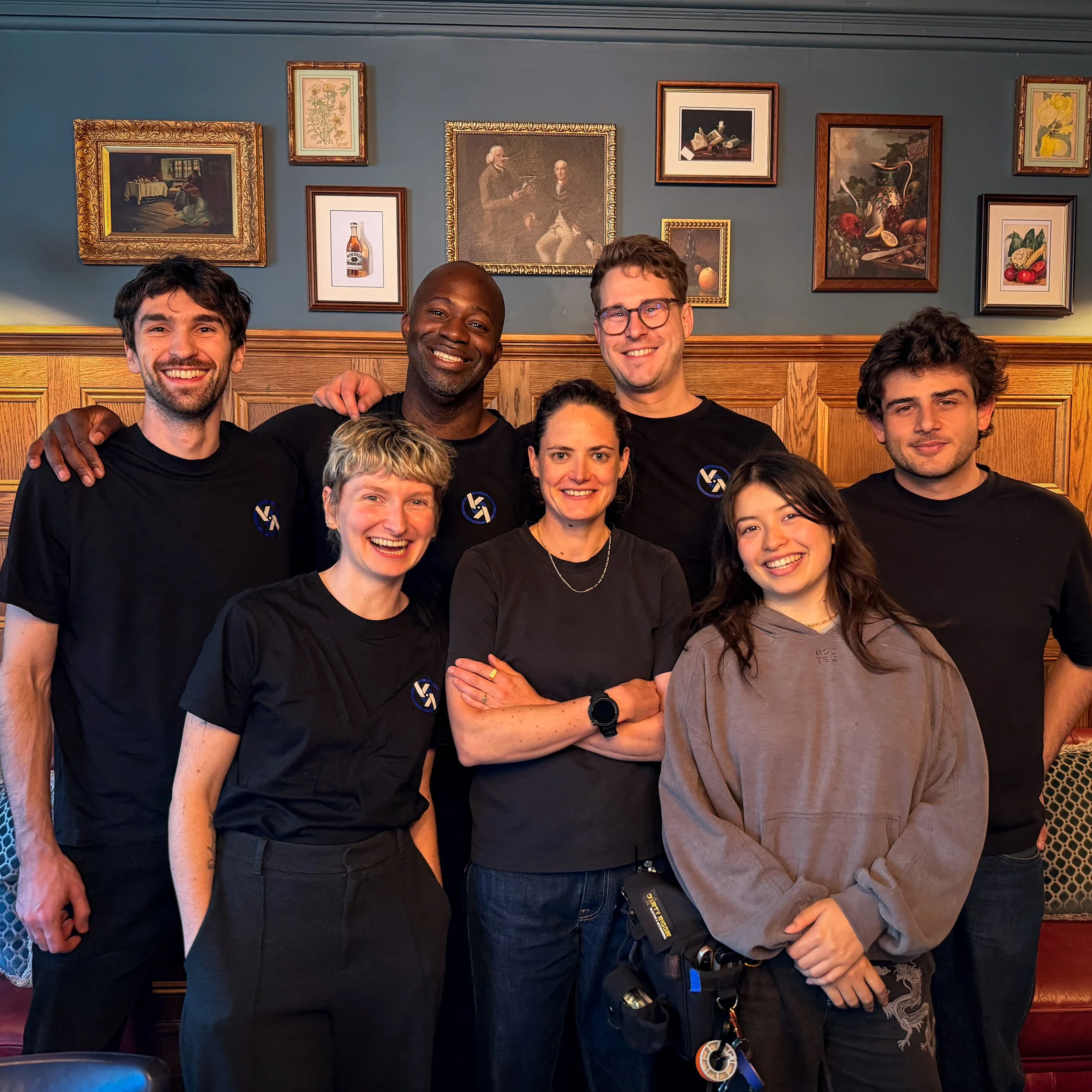 Group of seven people smiling and posing indoors in front of framed artwork on a blue wall, some wearing black shirts with a logo, others casual clothing.