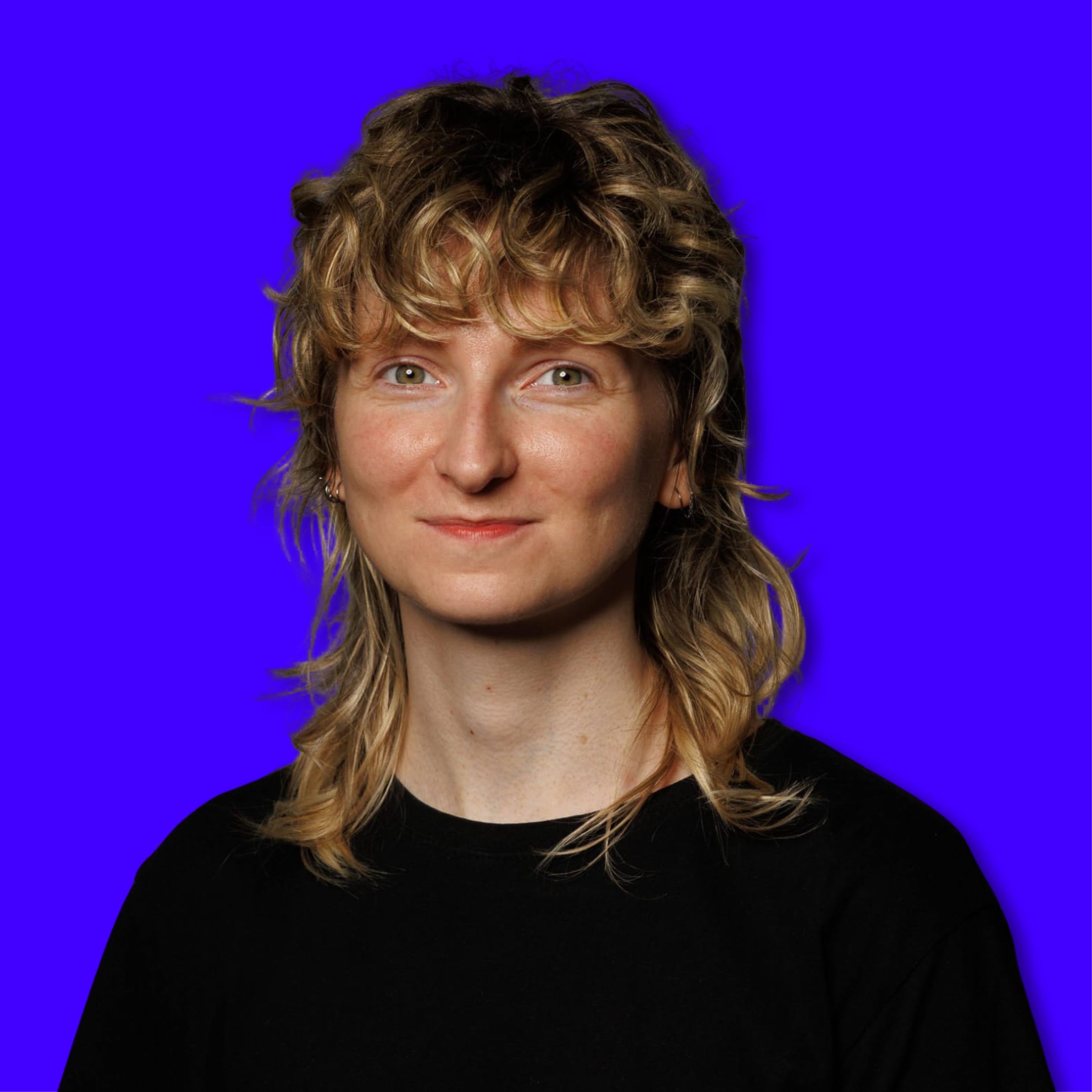 A woman with curly, shoulder-length blond hair and blue eyes, wearing a black top, smiles against a solid blue background.