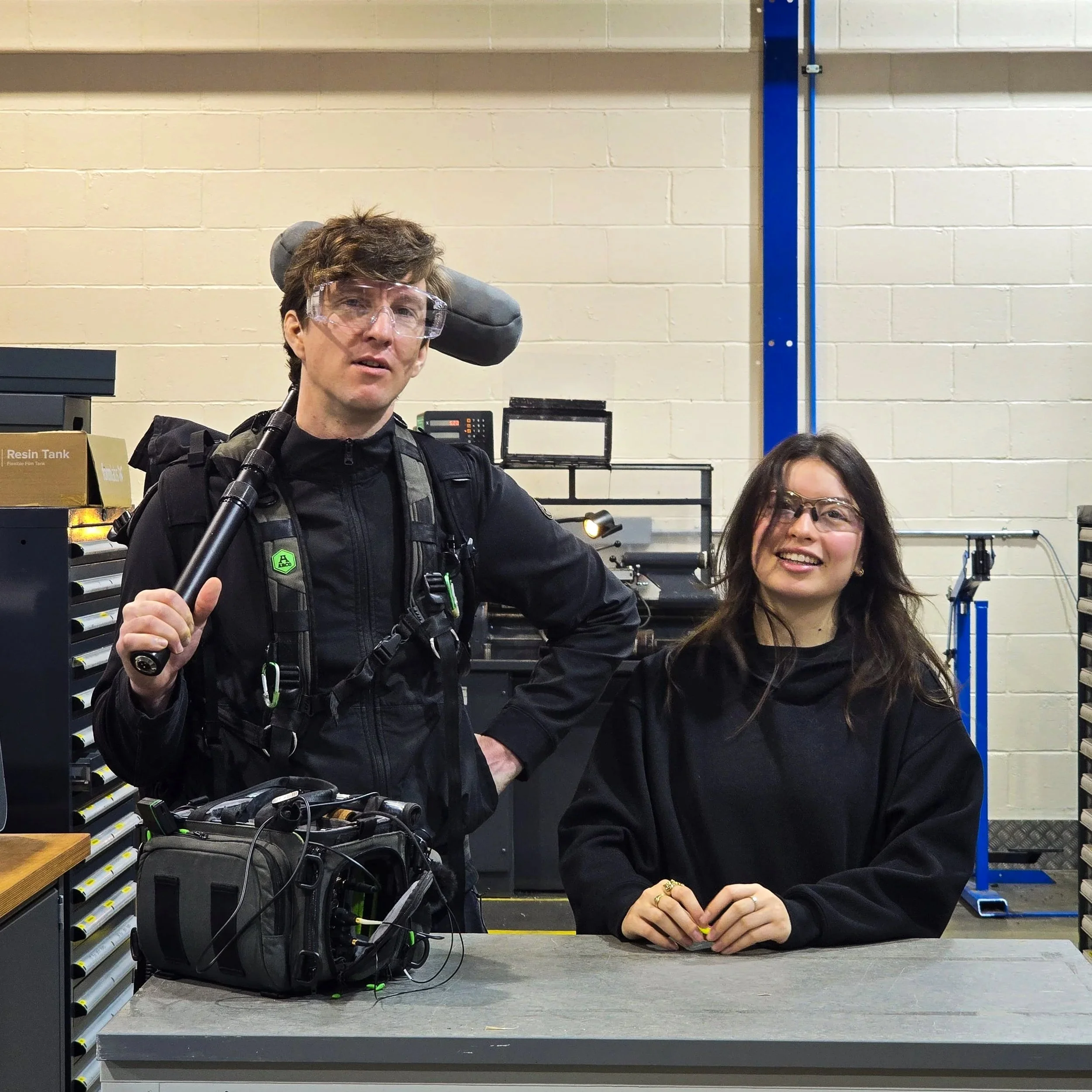 A man and a woman smiling and wearing safety glasses in an industrial workshop, with tools and equipment around them.