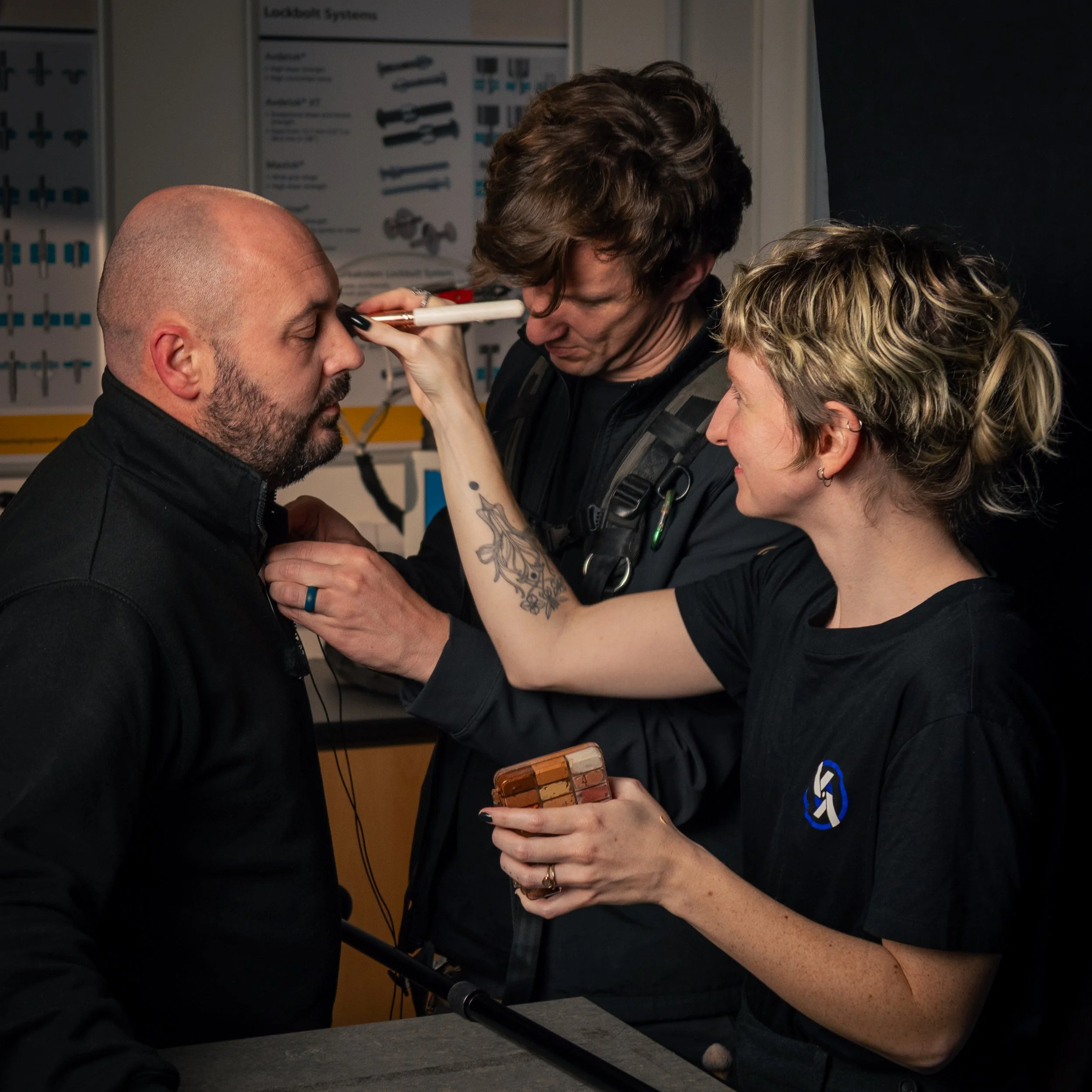 A makeup artist applies face paint to a man with a beard while a woman holds a makeup palette, with a makeup chart in the background.