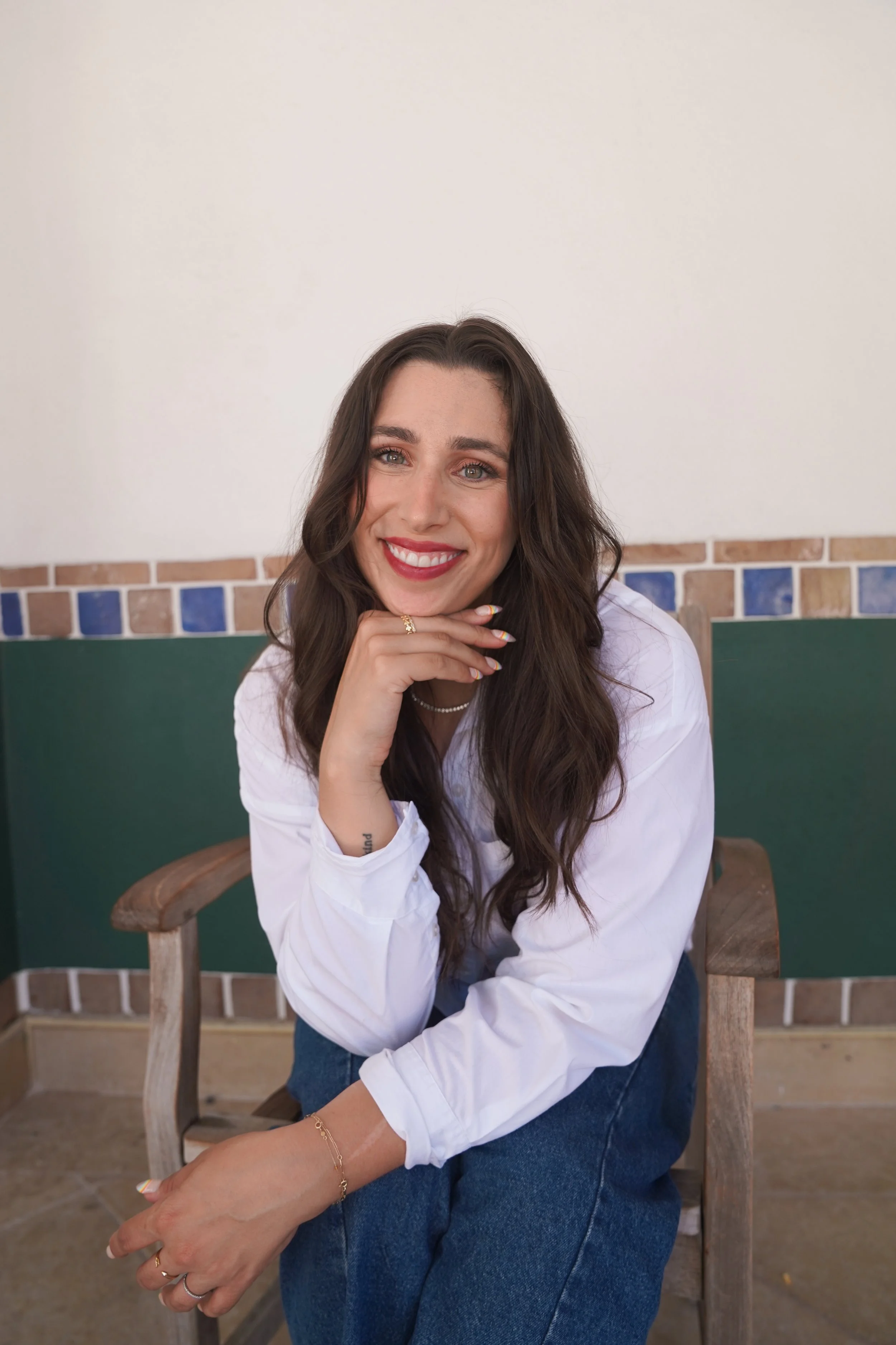 A woman with long wavy brown hair, smiling, seated on a wooden chair against a wall with blue and beige tile accents, wearing a white shirt and blue jeans.