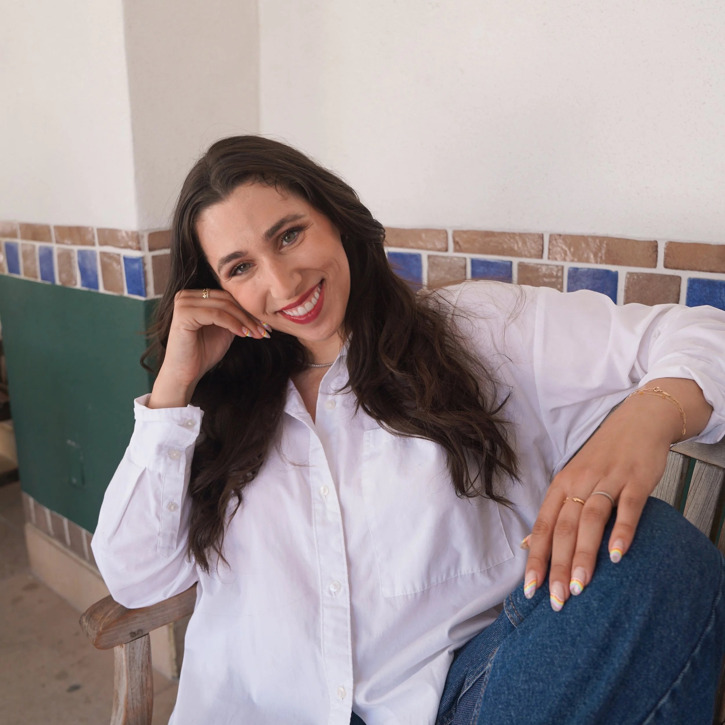 Smiling woman with long brown hair, wearing a white blouse, sitting on a wooden chair, with a brick and tile wall in the background.