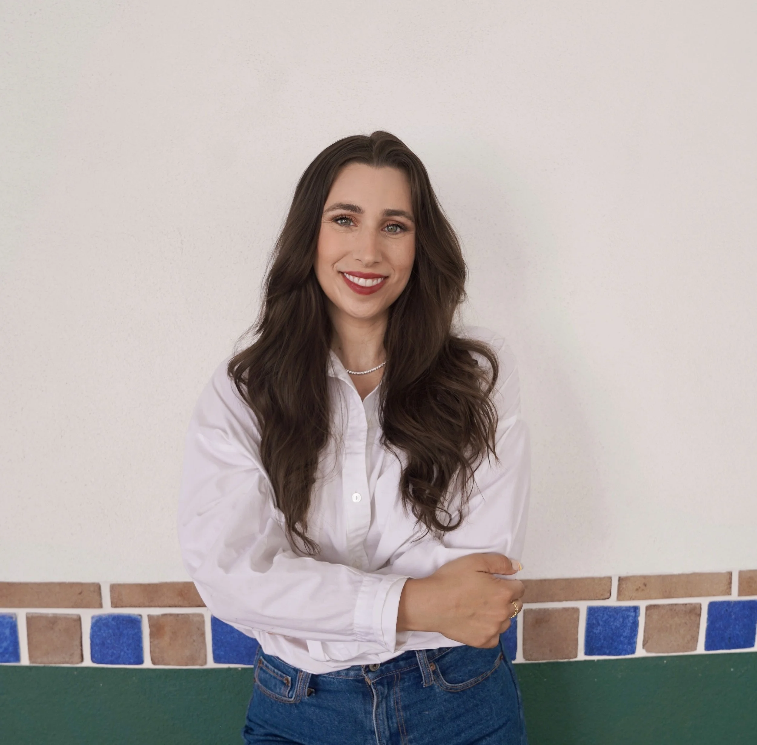 Smiling woman with long brown hair wearing a white blouse and blue jeans standing in front of a white wall with a green and blue tile border.