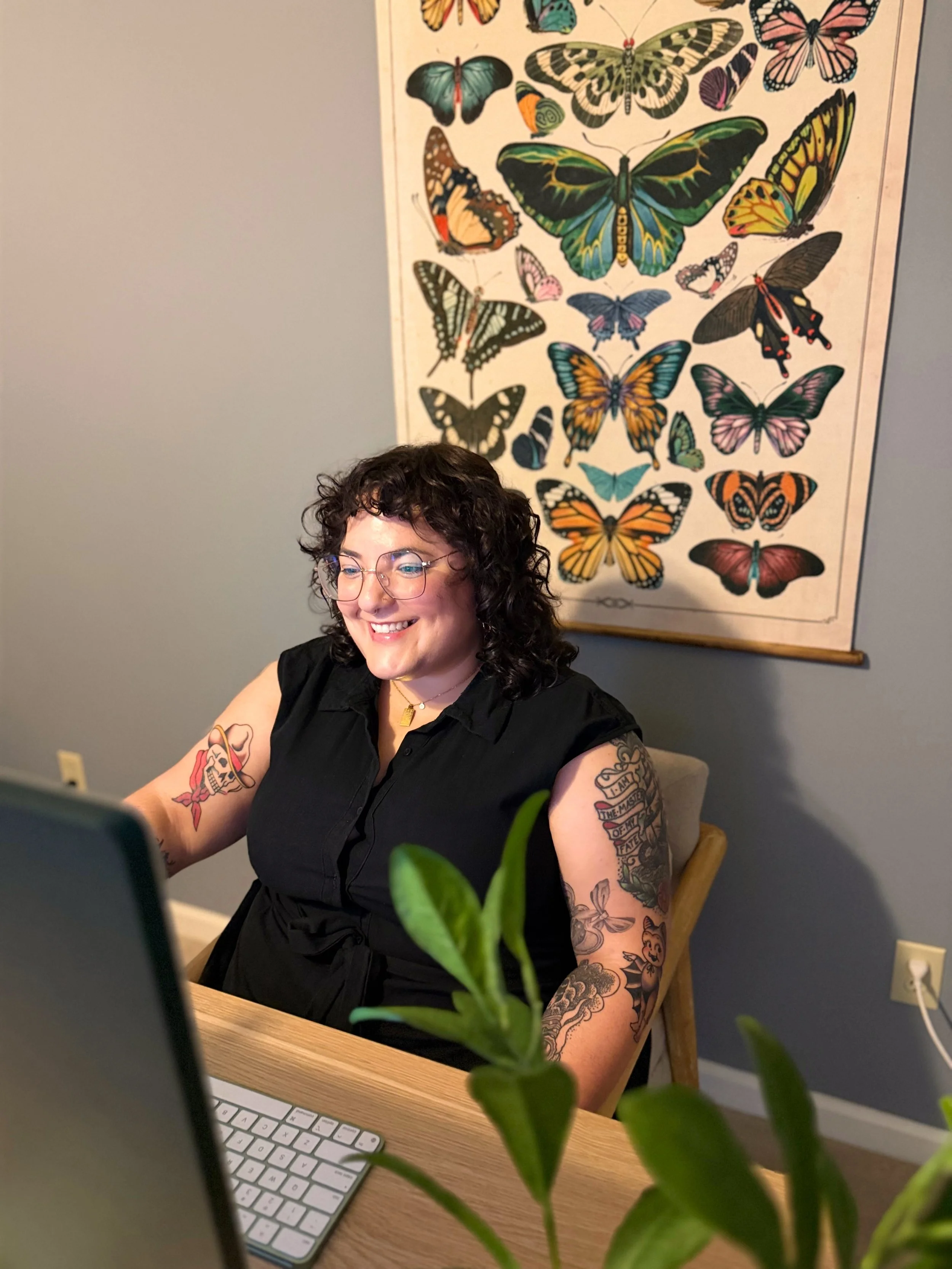A smiling woman with curly hair and tattoos on her arms working at a laptop in an office space, with a butterfly poster on the wall behind her and a green plant in the foreground.