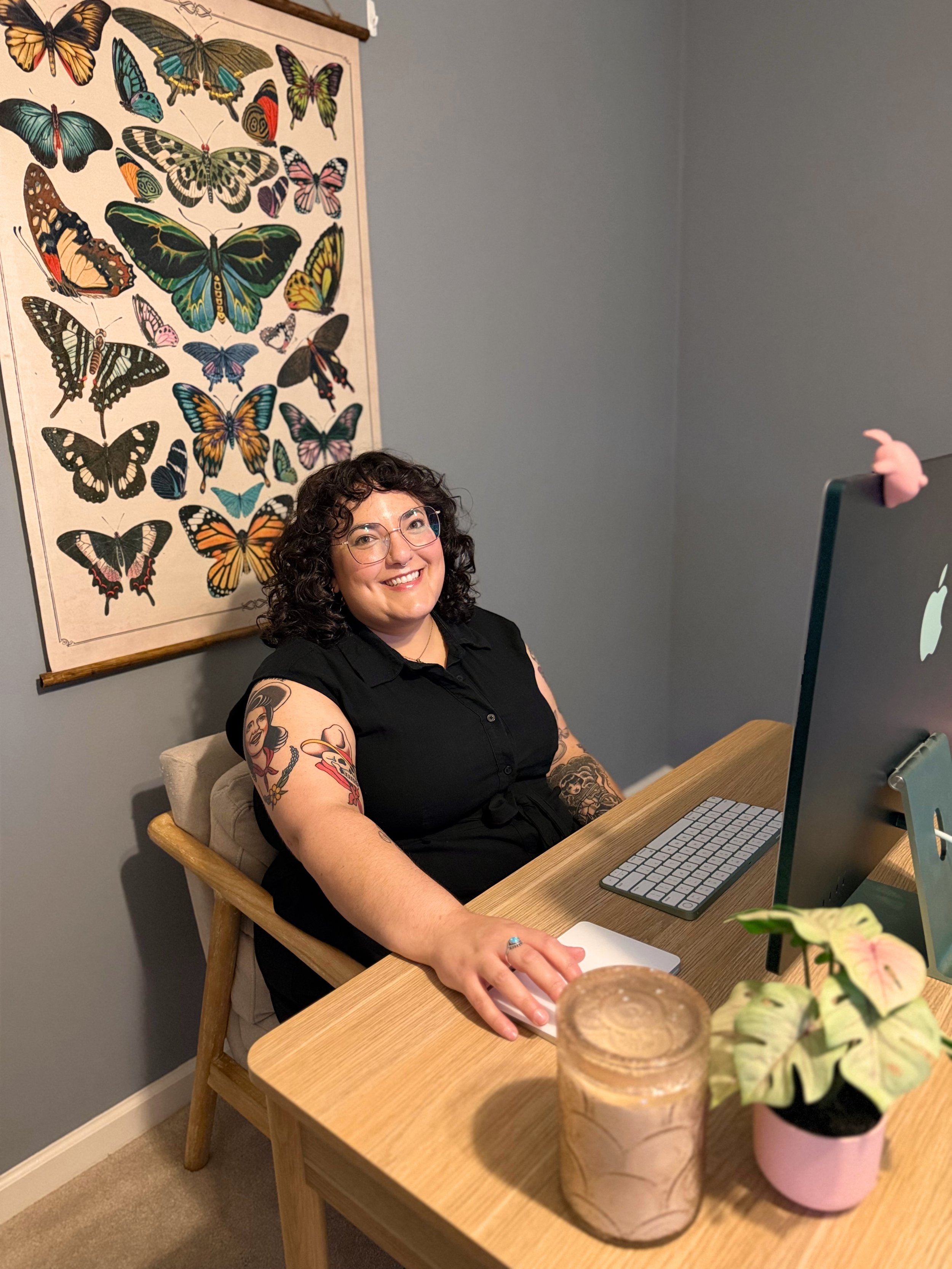 A woman with curly dark hair and glasses smiling at a desk with a computer screen. She has tattoos on her arms. Behind her is a poster of colorful butterflies on a wall.