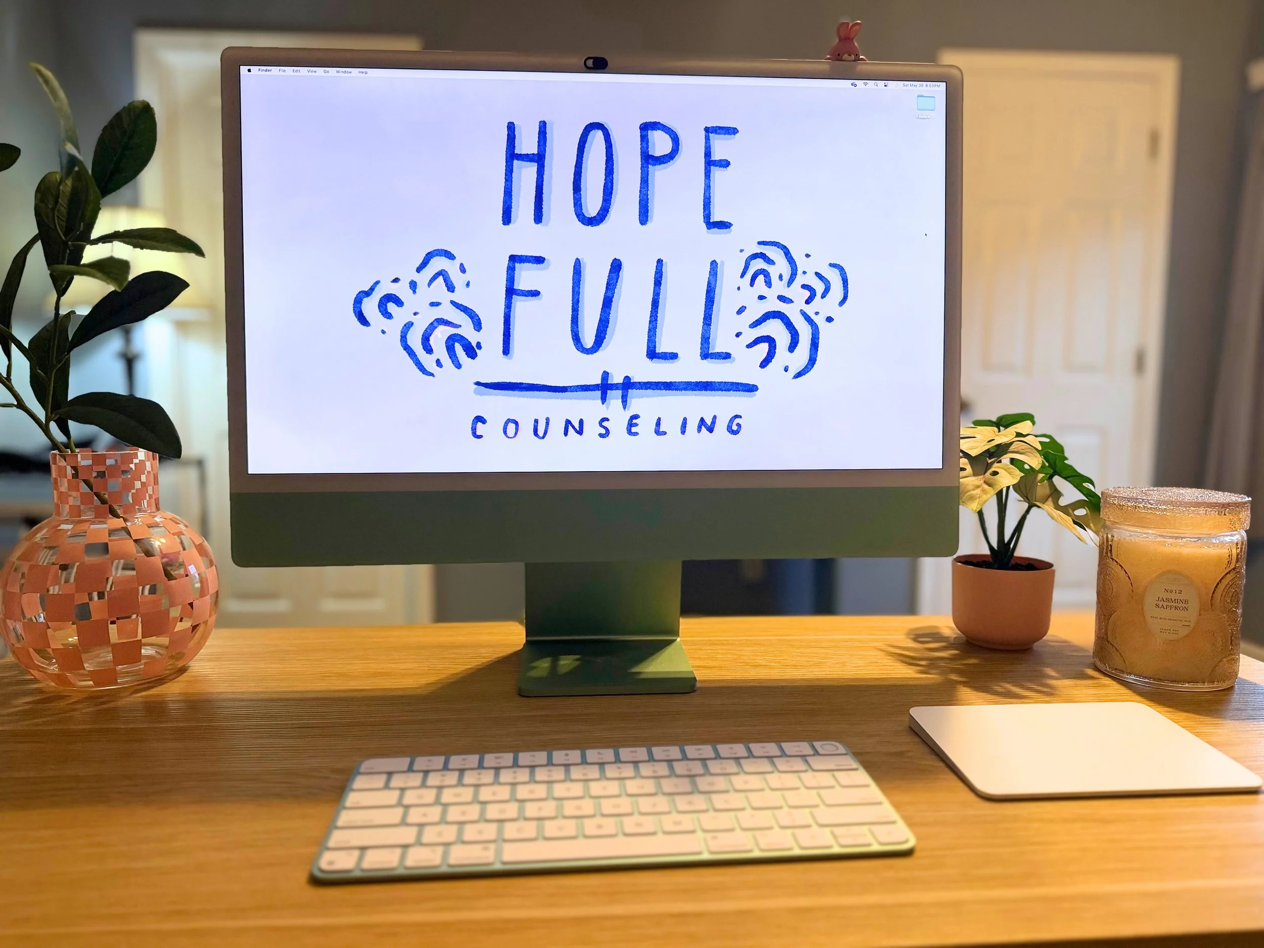 A computer monitor on a wooden desk displays a drawing with the words 'HOPE FULL' and 'COUNSELING'. The desk has a pink wicker-style vase with a plant, a purple container with a yellow flower, and a white keyboard and mouse. There is a small pink bunny figurine on top of the monitor. The background includes a plant and a doorway.