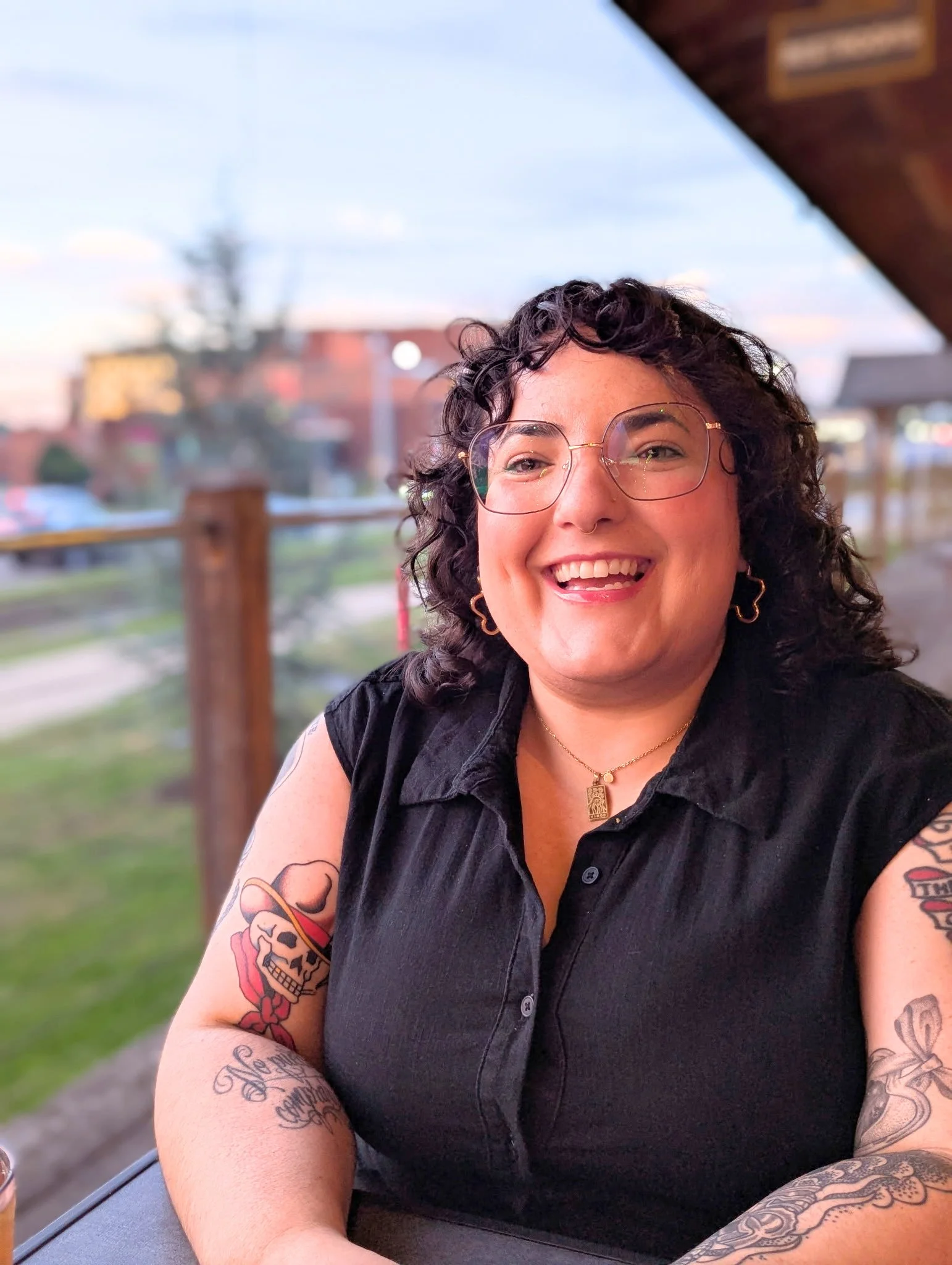 A woman with curly hair, glasses, and tattoos on her arms, smiling at the camera, sitting outdoors at sunset.