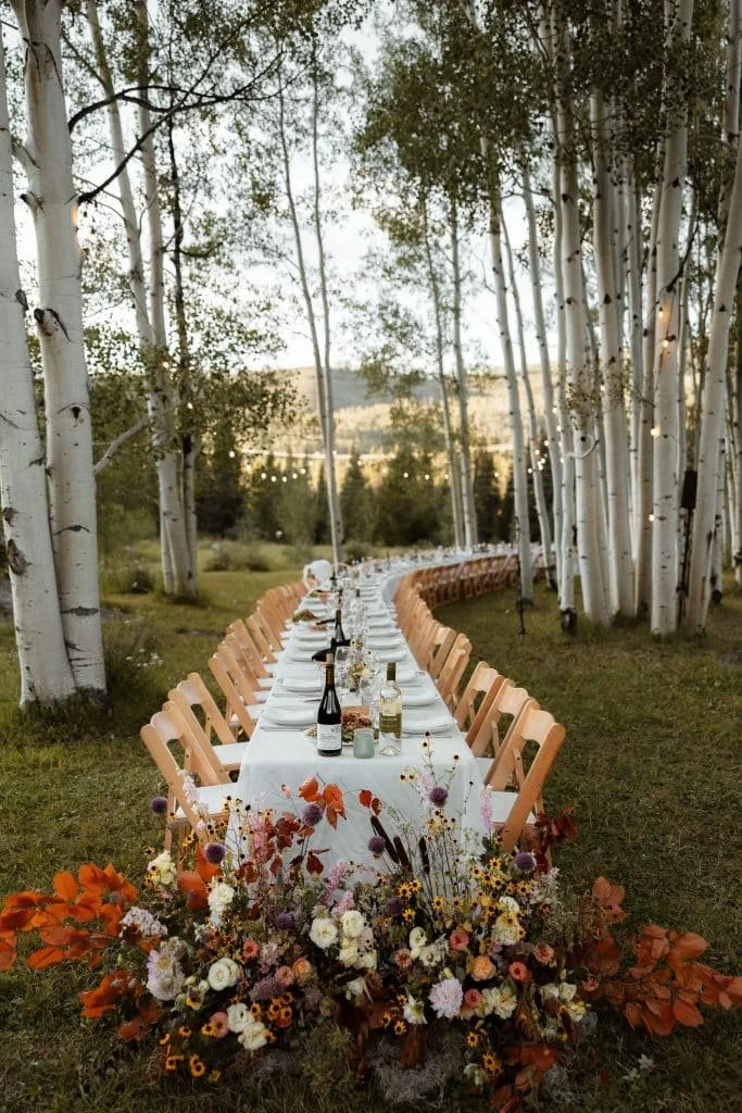 Outdoor wedding reception table setting at Beyul Retreat, with a long white-linen table, peach chairs, floral centerpieces, and string lights strung through a forest of white birch trees.