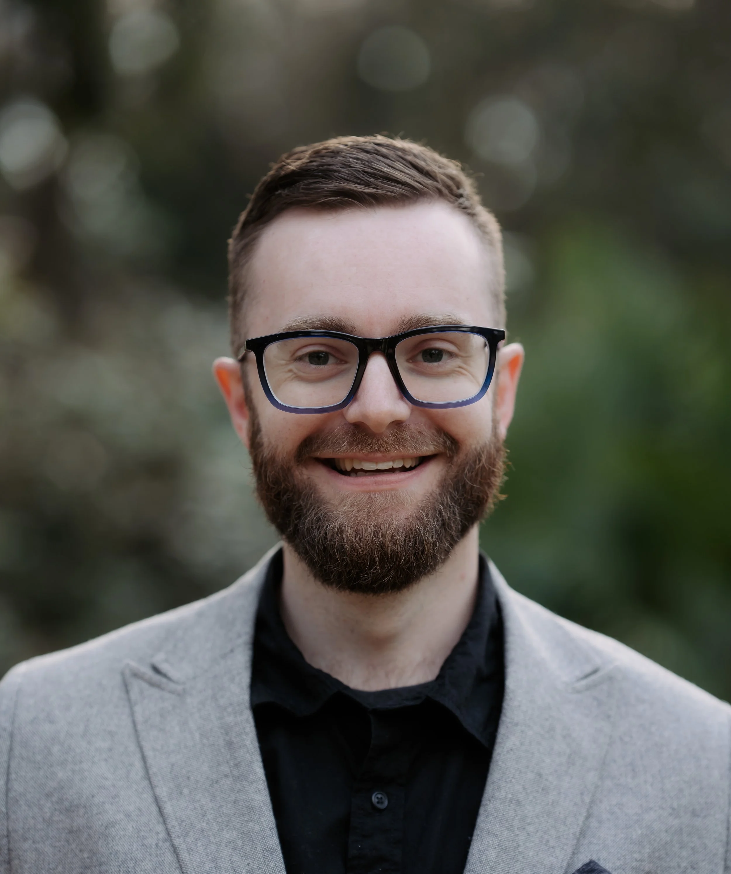 A smiling man with glasses, a beard, and a short hairstyle wearing a gray blazer and black shirt outdoors.