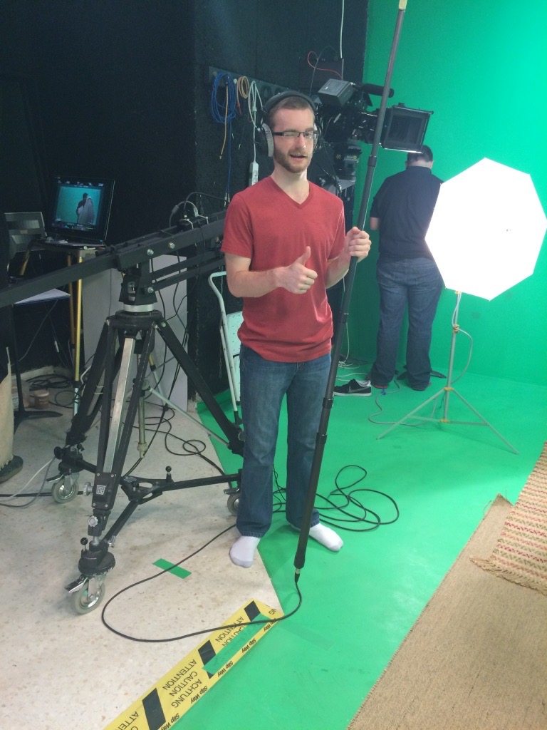 Man in a red shirt and jeans giving a thumbs-up in a green screen photo or video studio with professional lighting and filming equipment.