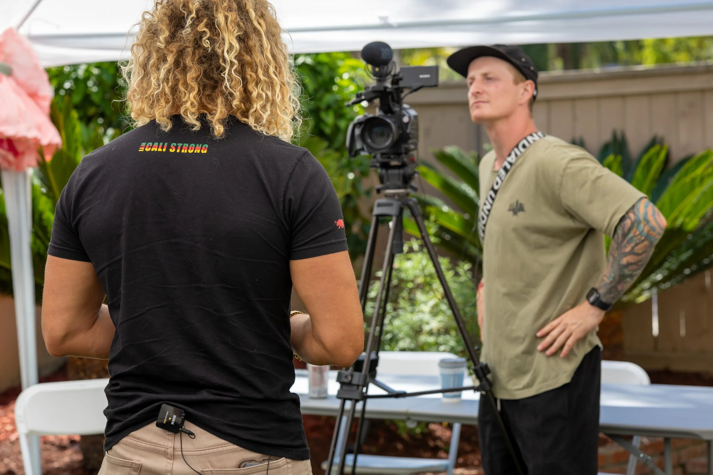 A man with tattoos wearing a beige t-shirt and a black cap is standing outdoors with his hands on his hips, being filmed by a camera operator with curly blonde hair wearing a black t-shirt that says 'Calif. Strong'. The scene is set in a backyard wit