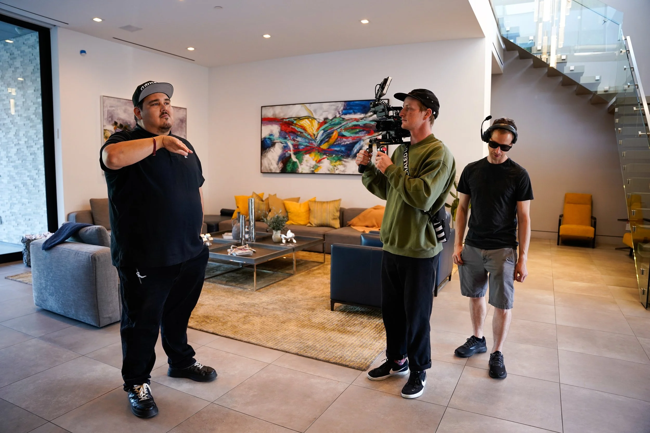 Three men involved in a filming or photography project inside a modern living room. One man appears to be giving directions, while another operates a camera on a shoulder rig, and the third looks on. The room has a colorful abstract painting on the w