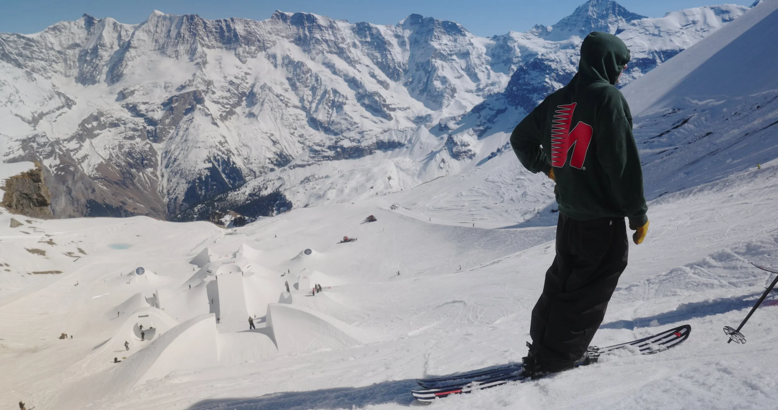 ski athlete standing above massive sculpted snow feature during Swatch Nines Switzerland, documented for snowboard event production and freeski documentary coverage.
