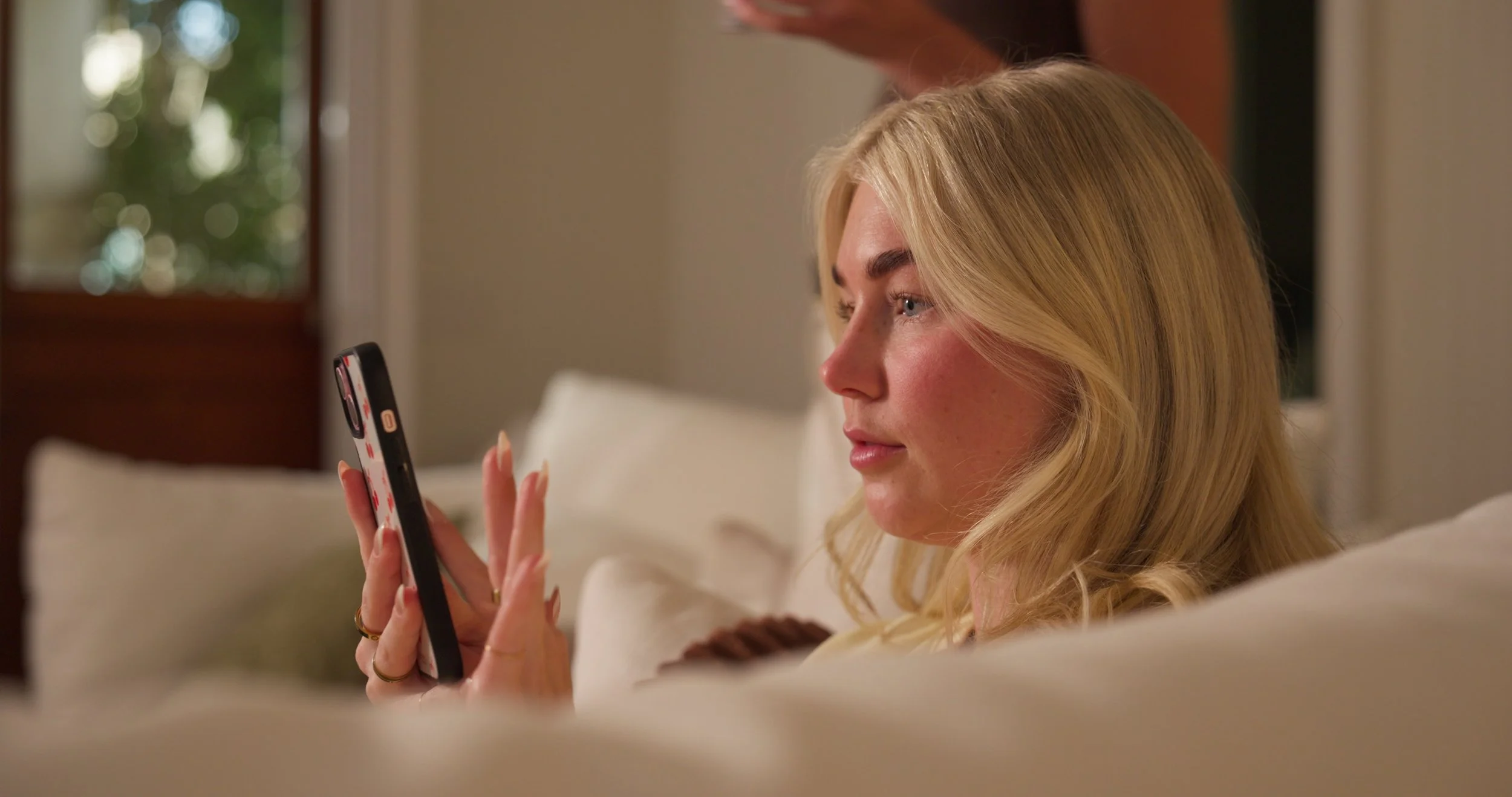 A blonde woman relaxing on a bed, looking at her smartphone in a cozy room with a window in the background.