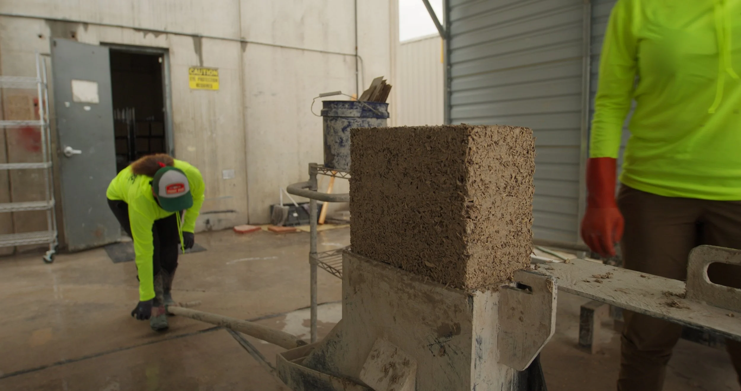 Construction workers wearing neon green shirts and safety gloves working on a construction site with a brick and concrete block in the foreground.