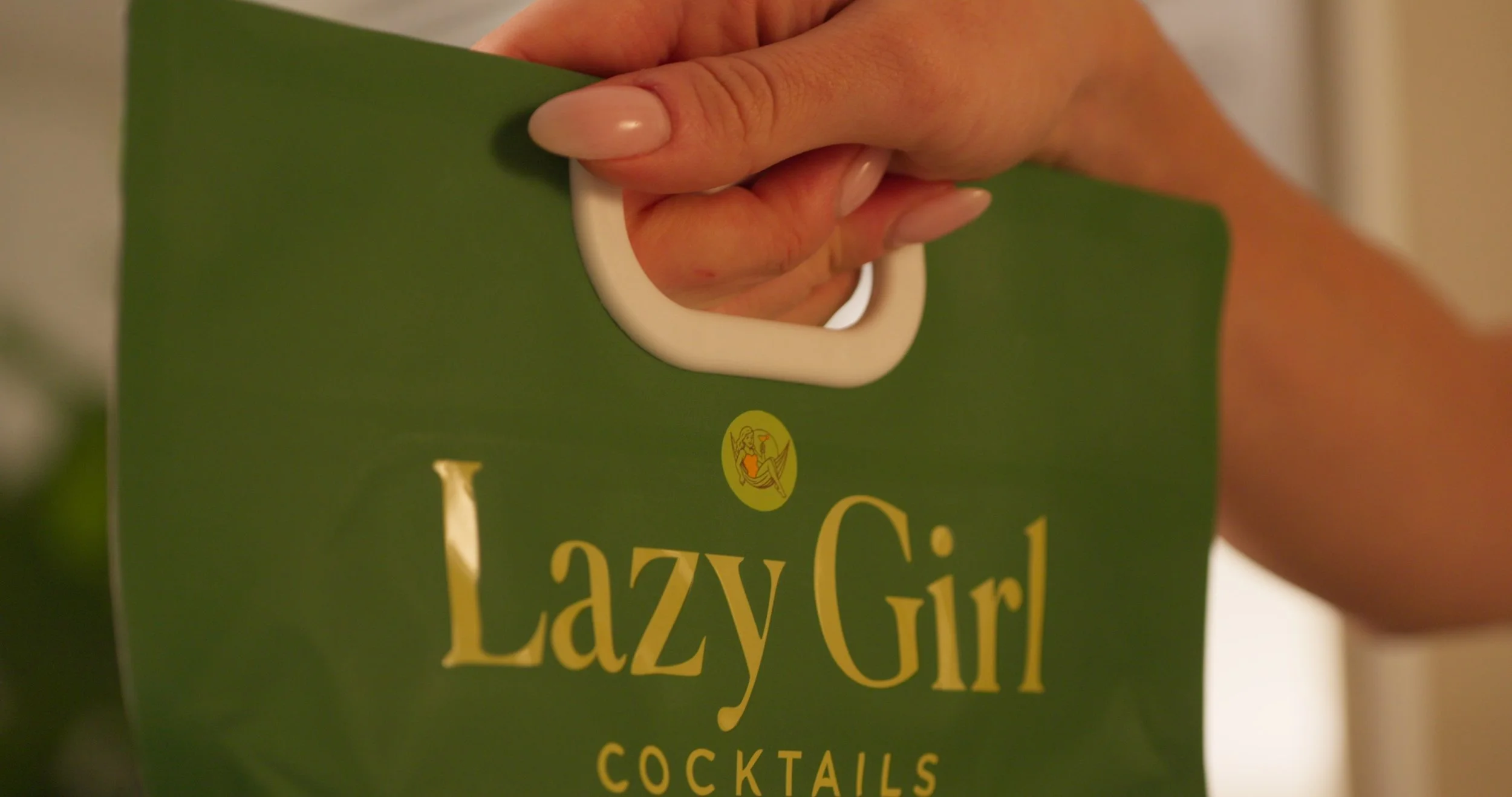 Close-up of a person's hand holding a green bag labeled 'Lazy Girl Cocktails', with a white handle.